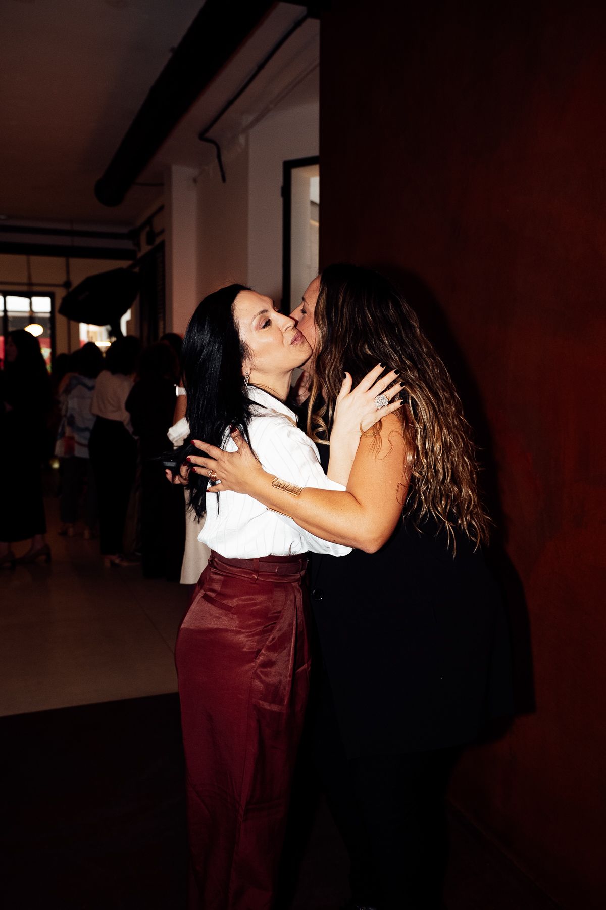 Two women sharing a warm embrace and kiss at a dimly lit social event.