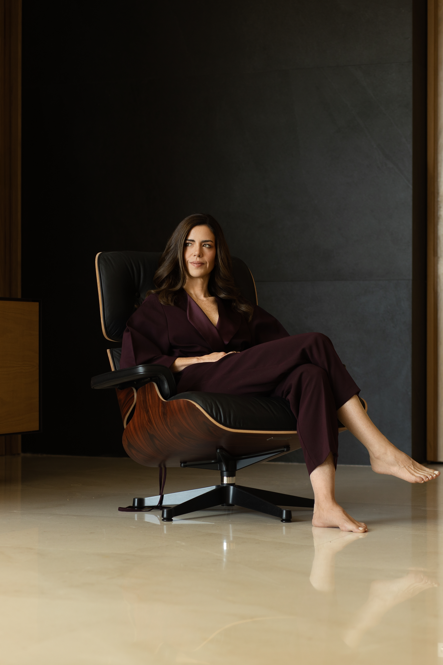 Woman in burgundy suit relaxing barefoot in classic Eames lounge chair against dark wall on marble floor.