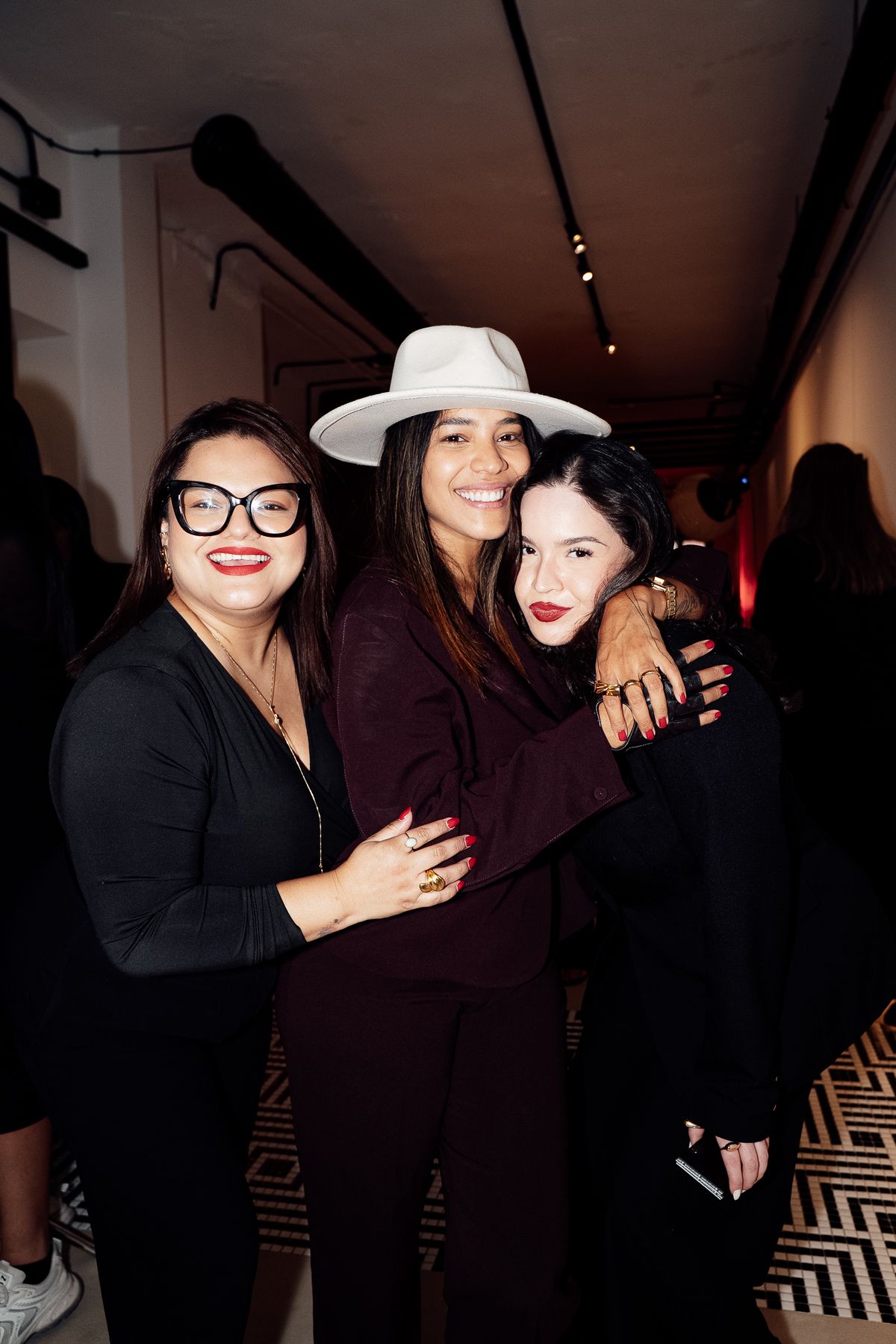 Three women pose together smiling at an indoor event, one wearing a white wide-brim hat and burgundy suit.