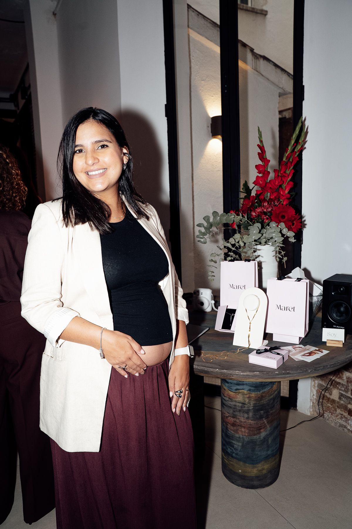 Smiling pregnant woman in white blazer and burgundy skirt posing at a jewelry brand event.