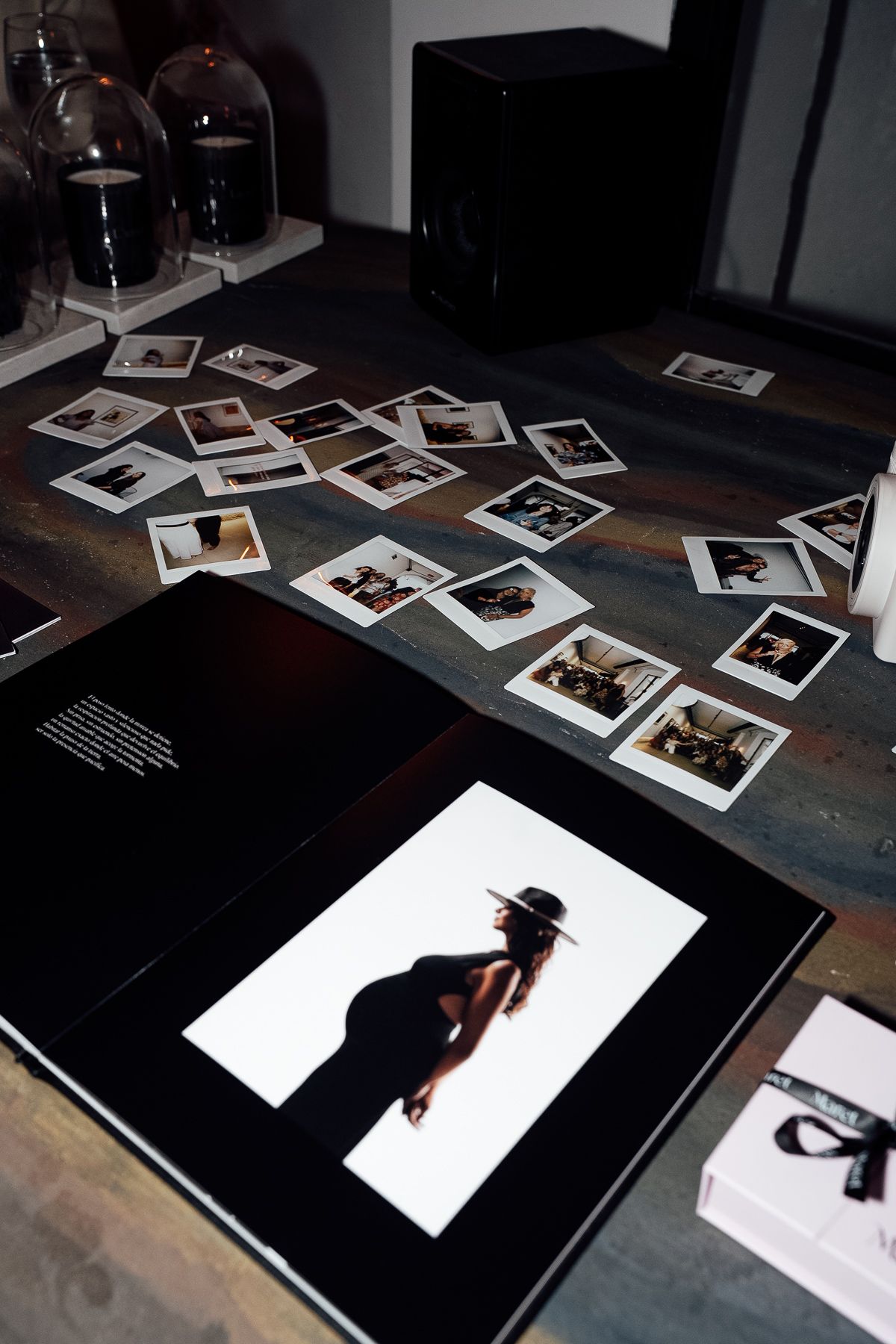 Open photo book with black cover displaying a woman in a hat, surrounded by scattered Polaroid prints on a dark wooden table.