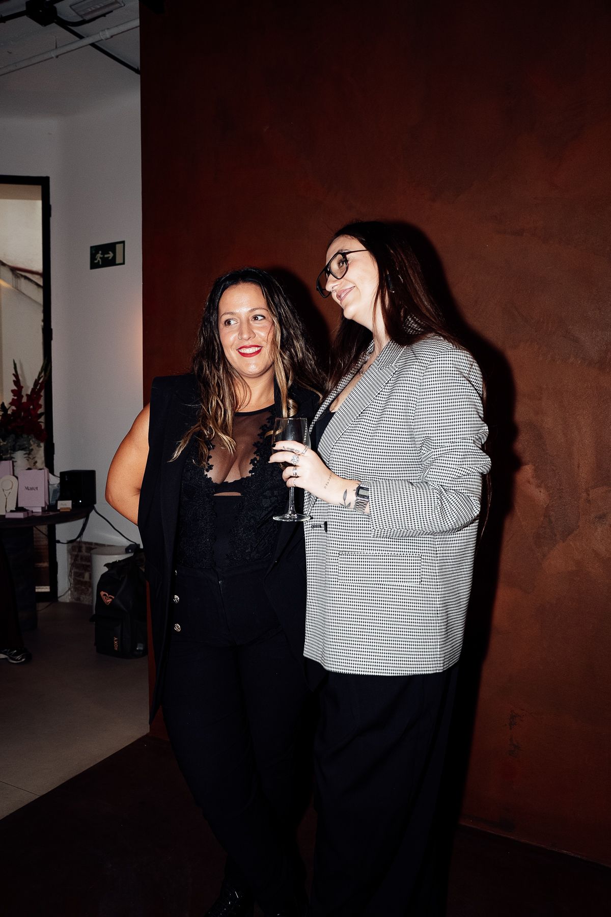 Two women smiling at an indoor event, one in black outfit and one in gray blazer holding a champagne glass.