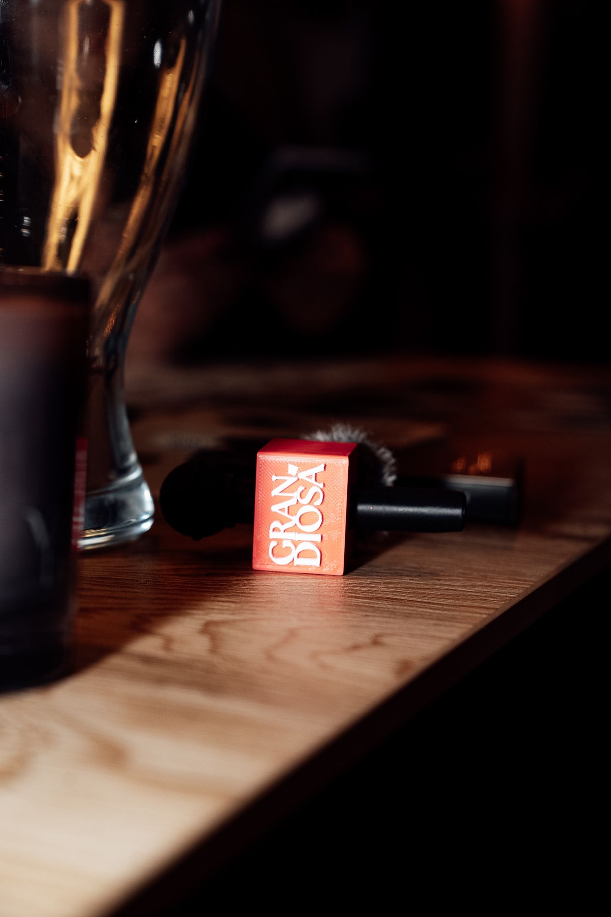 Red Gran Diosa perfume box on wooden surface with makeup brush and glassware in moody lighting.