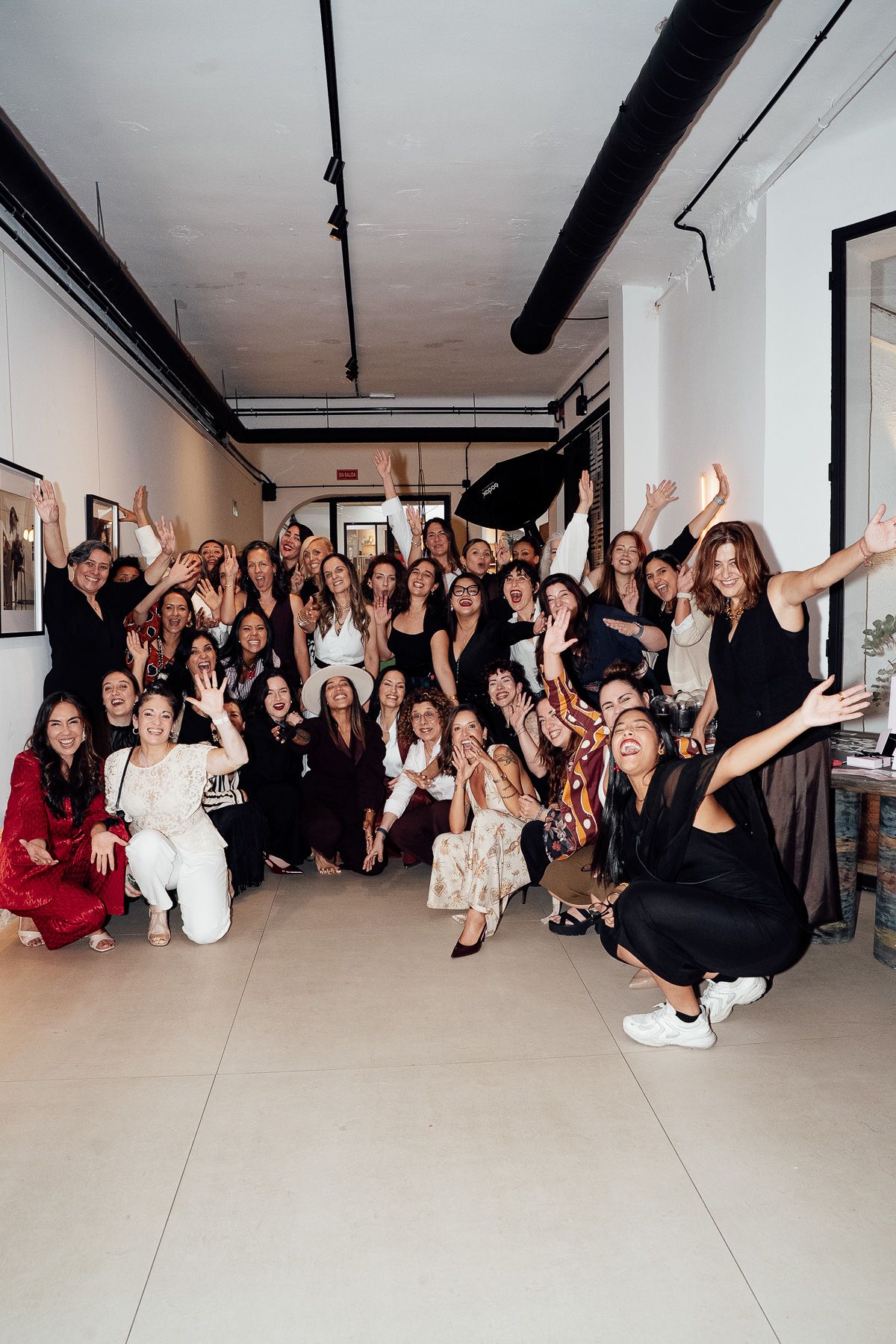 A large group of diverse women celebrating together in a modern gallery space, arms raised in excitement.