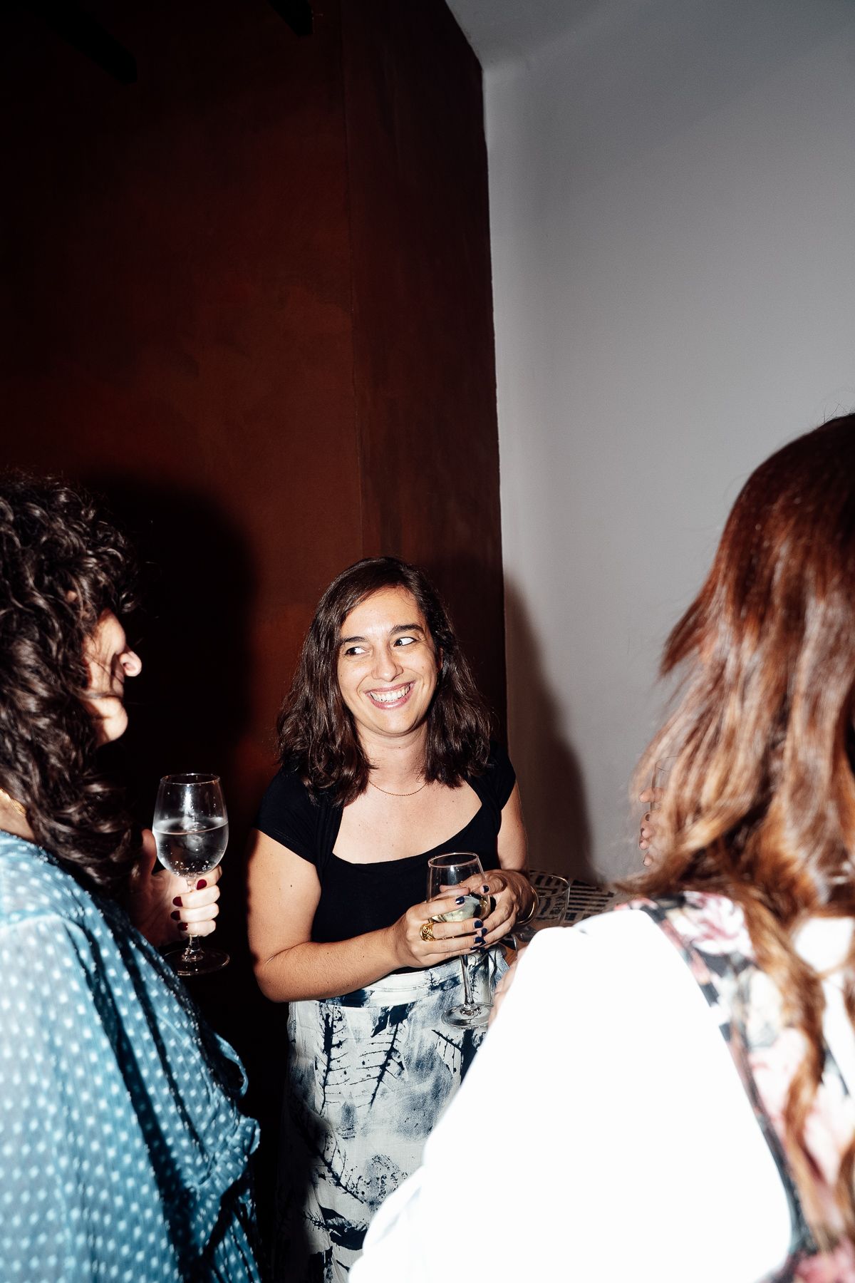 Woman smiling and holding wine glass at social gathering, conversing with friends indoors.