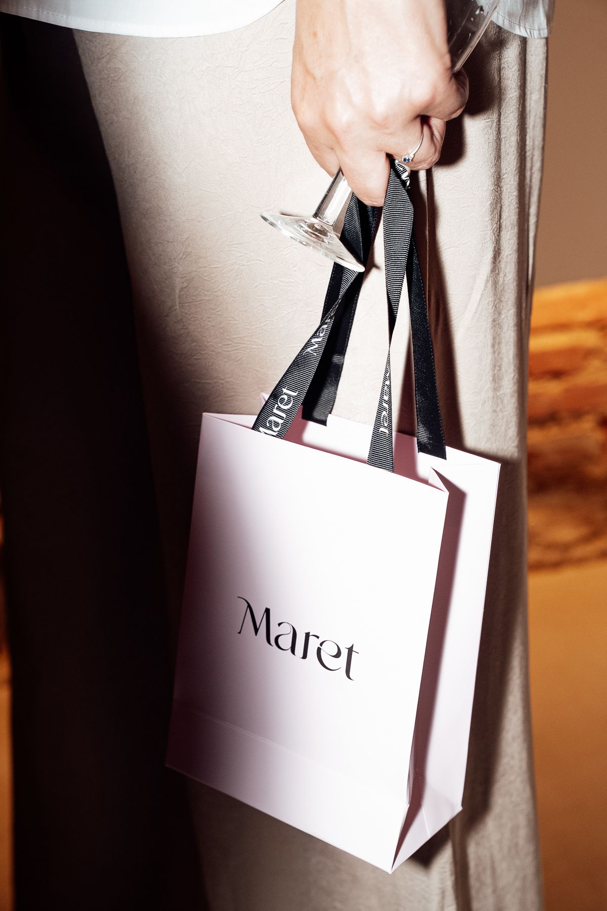 Hand holding a Maret branded shopping bag with black ribbon handles against a neutral background.