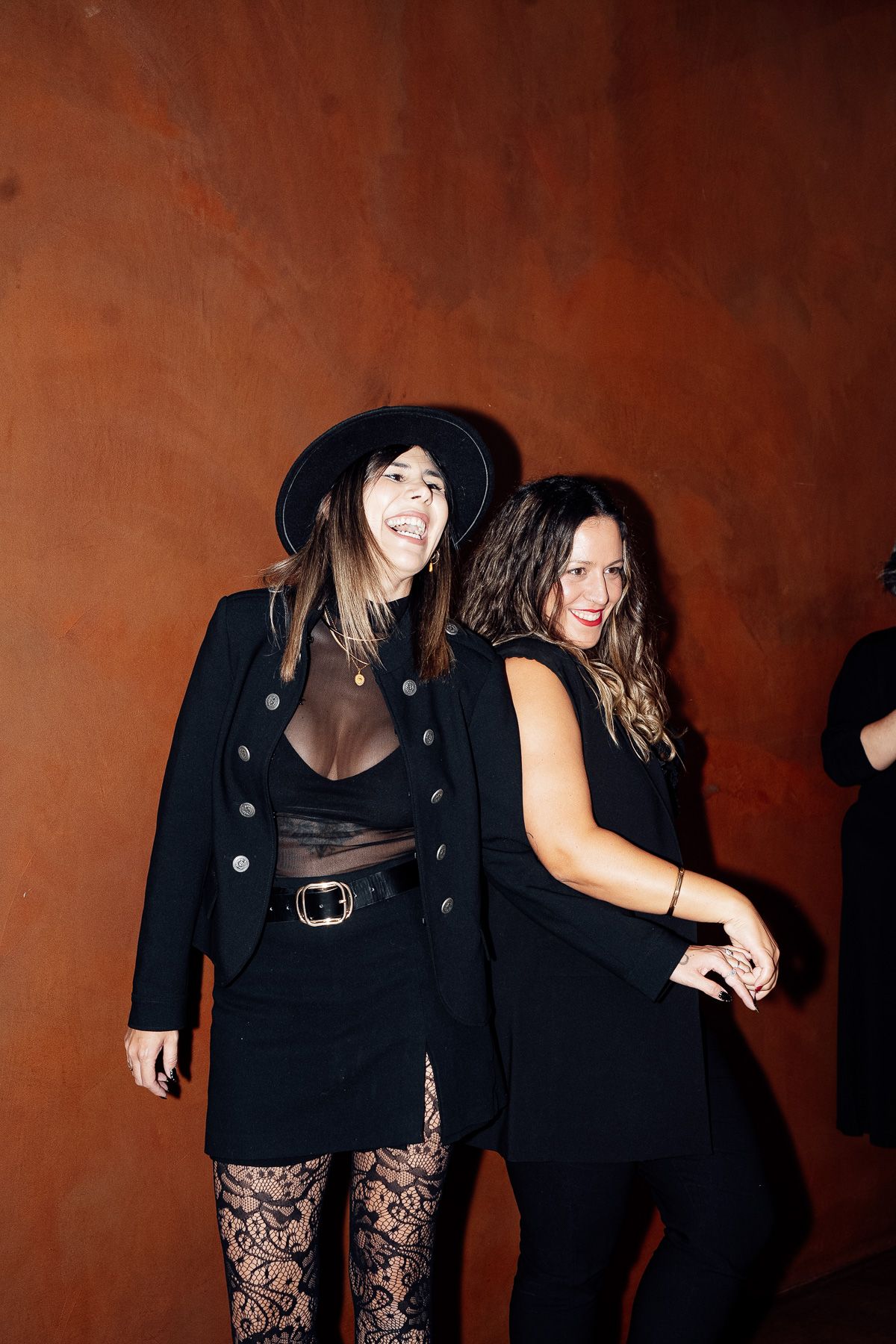 Two women in chic all-black outfits posing and smiling against a rustic brown wall at a fashion event.