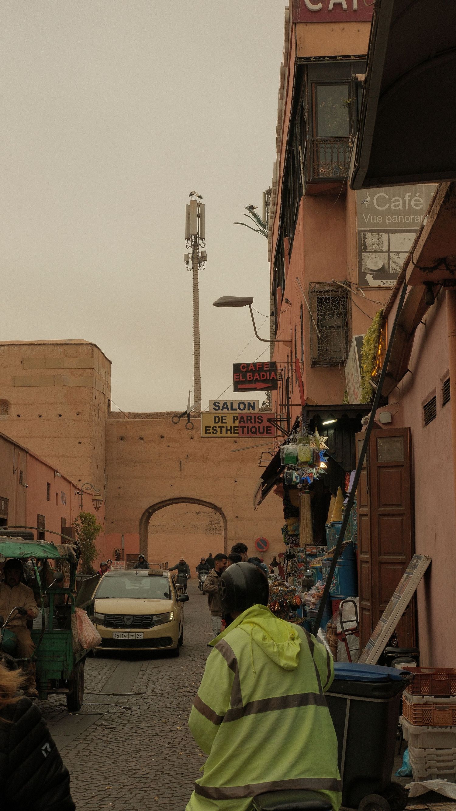Busy Marrakech medina street with a worker in yellow vest, pink buildings, salon sign, and ancient city gate in background.