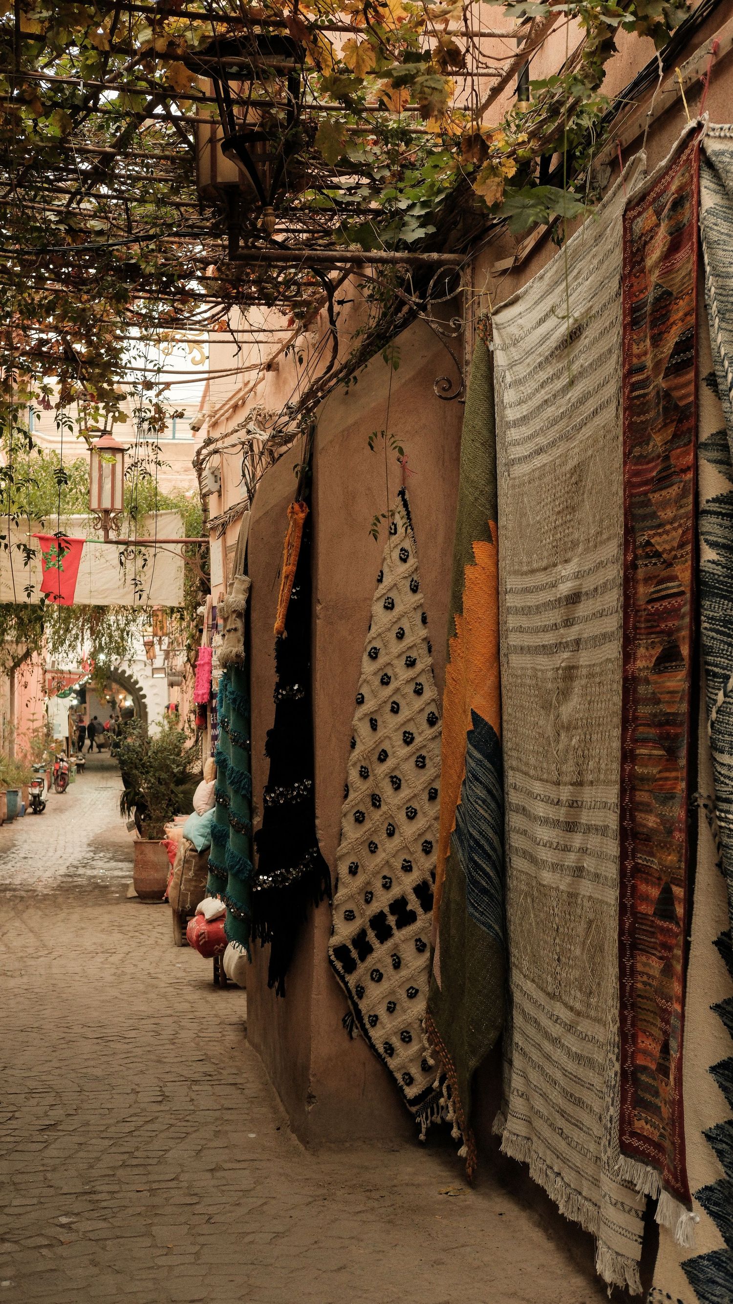Colorful rugs and textiles hanging on walls of a narrow cobblestone alley in a traditional Moroccan medina marketplace.