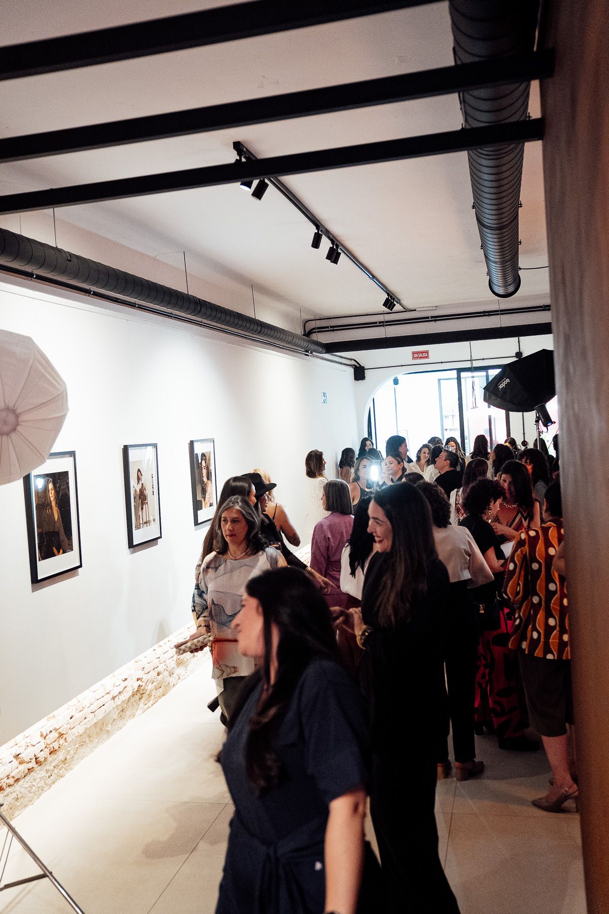Guests browse framed artwork at a crowded art gallery opening with white walls and track lighting.