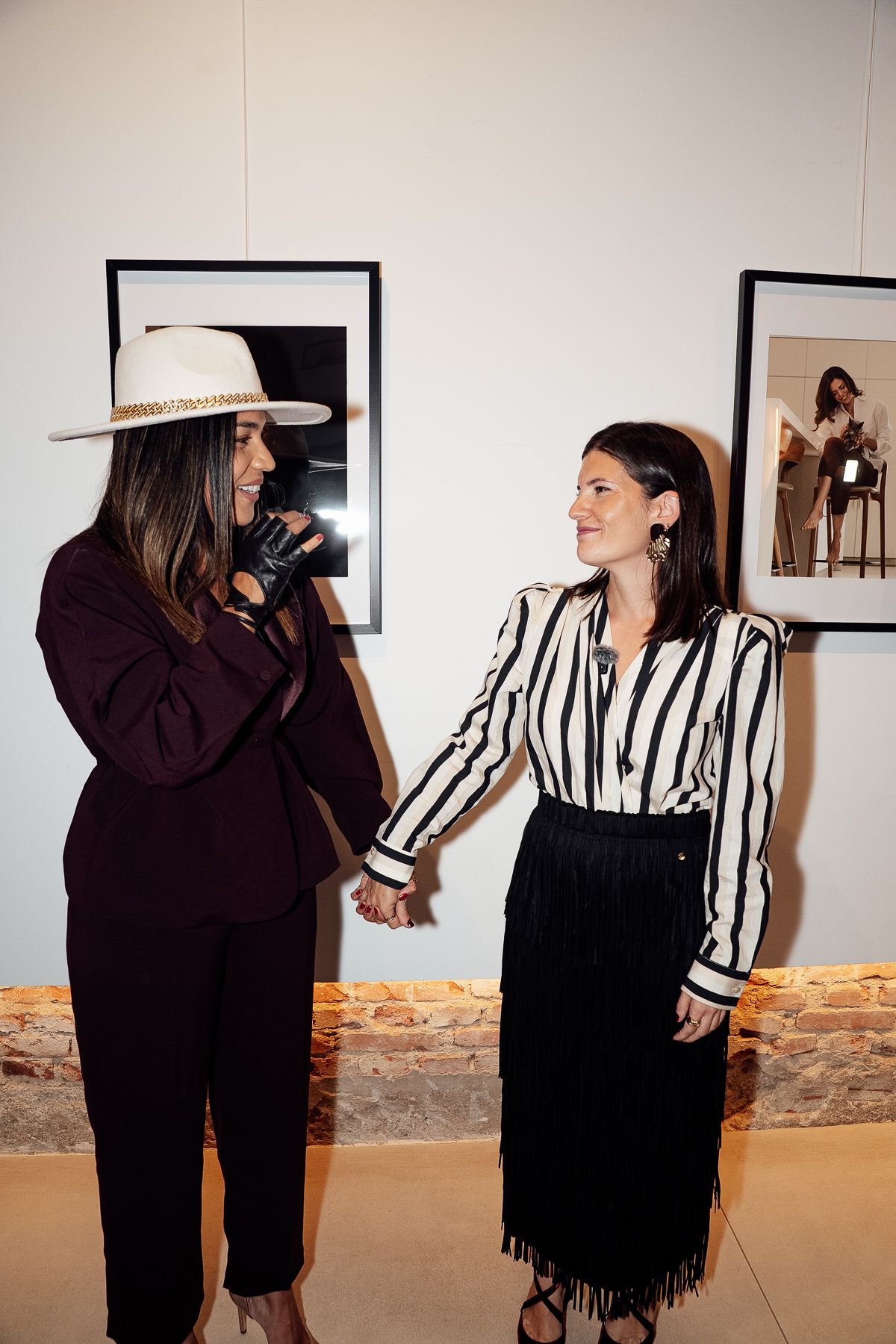 Two stylish women holding hands at an art gallery, one in a white hat and dark suit, the other in a striped blouse.
