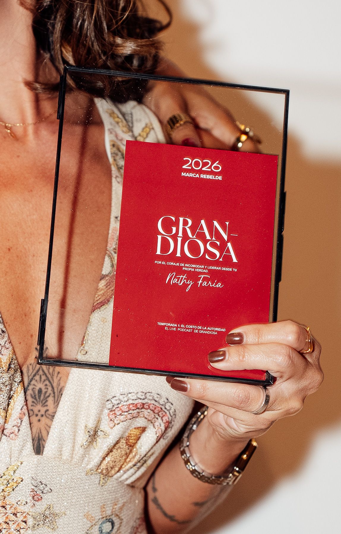 Woman holding a red Gran Diosa 2026 Marca Rebelde award plaque by Nathy Faria, showcasing tattoos and jewelry.