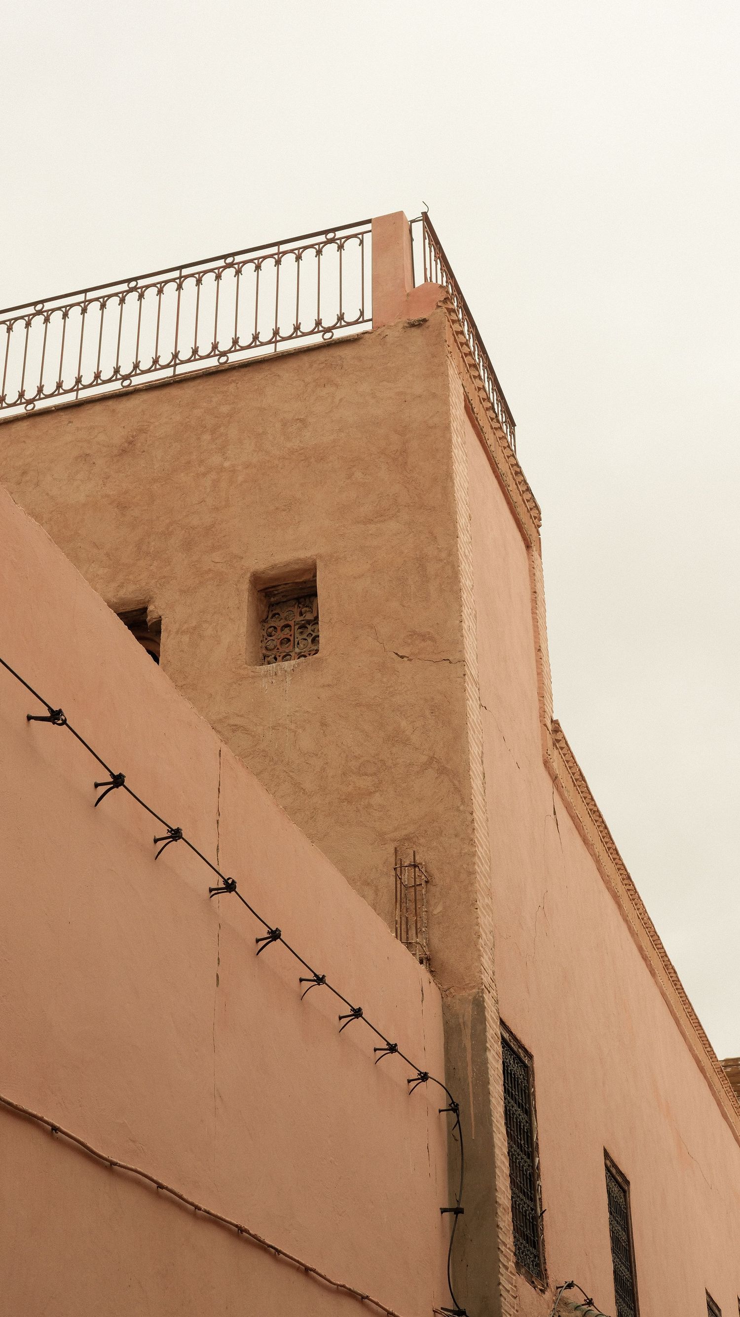 A low-angle view of a terracotta adobe building with a rooftop railing and barbed wire fence against an overcast sky.