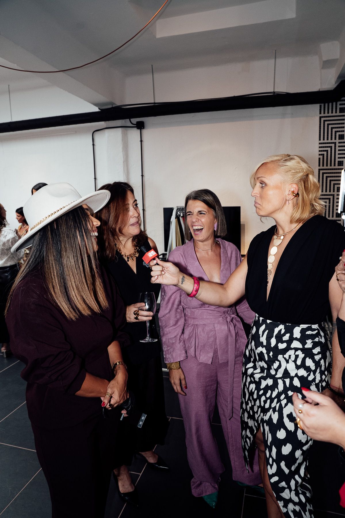 Four women socializing and laughing at an upscale event, holding drinks in a modern interior space.