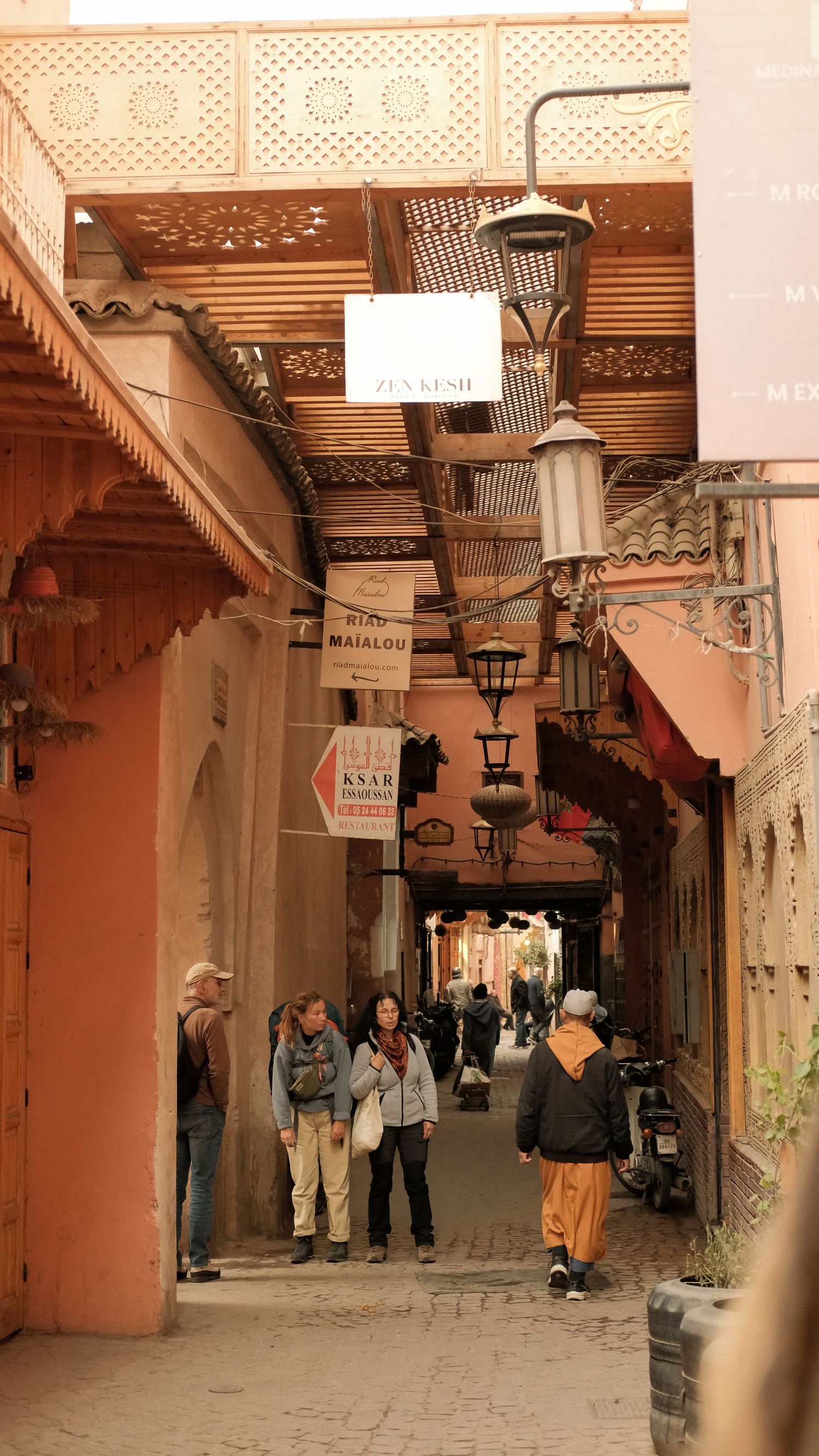 Narrow alleyway in Marrakech medina with tourists, traditional lanterns, and shop signs in warm terracotta tones.