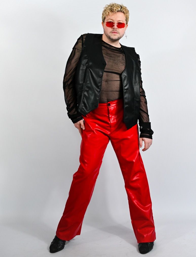 Stylish man in red vinyl pants, black mesh top, leather jacket, and red sunglasses posing boldly.