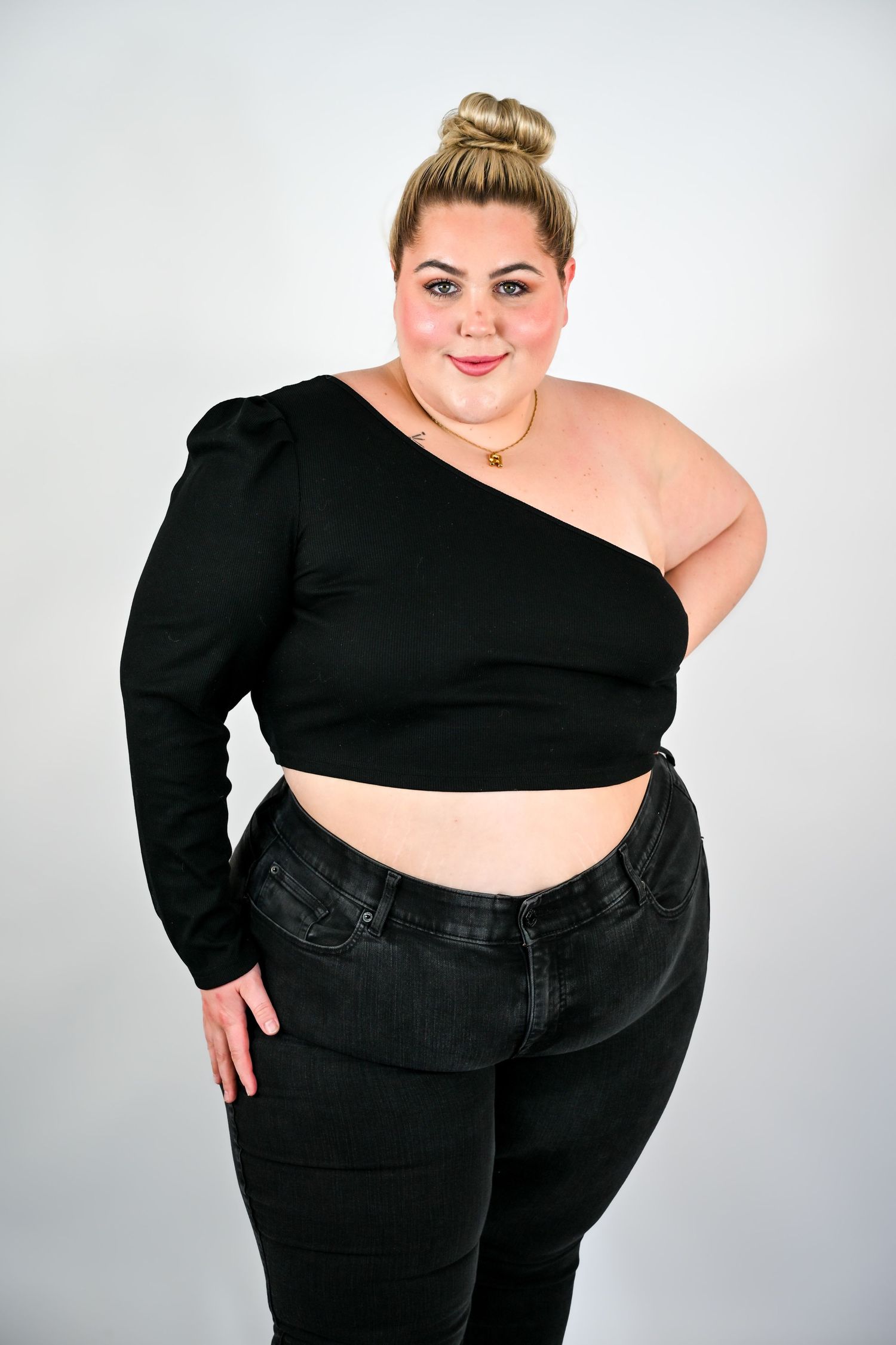 Plus-size blonde woman in black one-shoulder crop top and dark jeans posing against gray background.