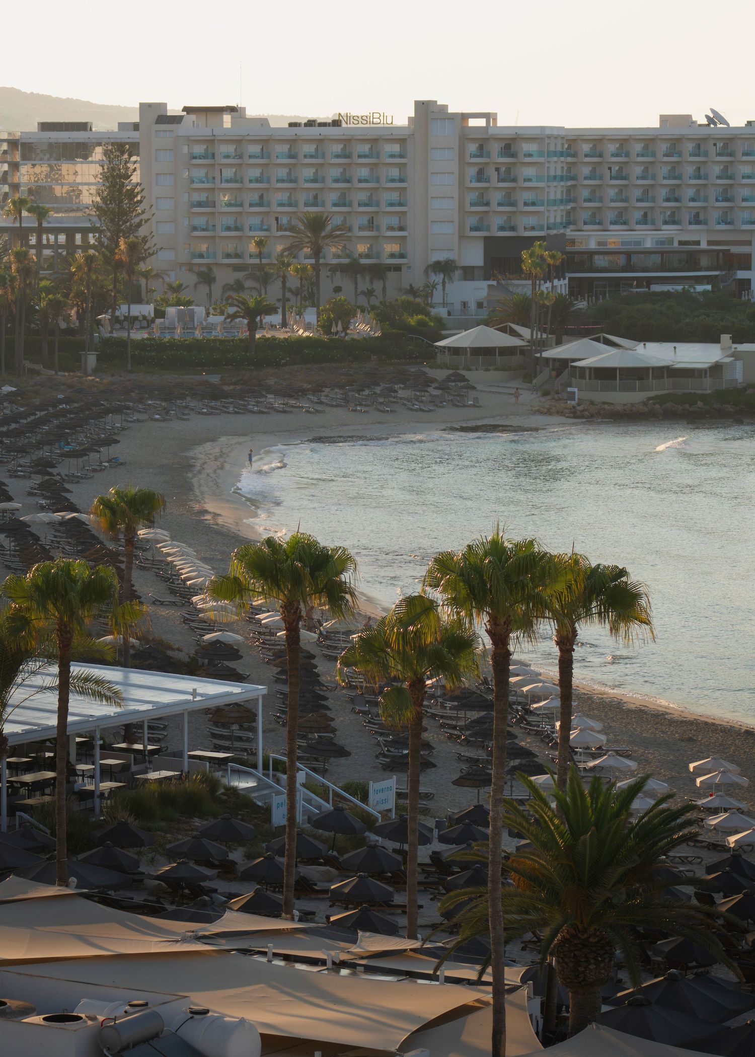 Aerial view of a Mediterranean beach resort with palm trees, sun loungers, and a large hotel at sunset.