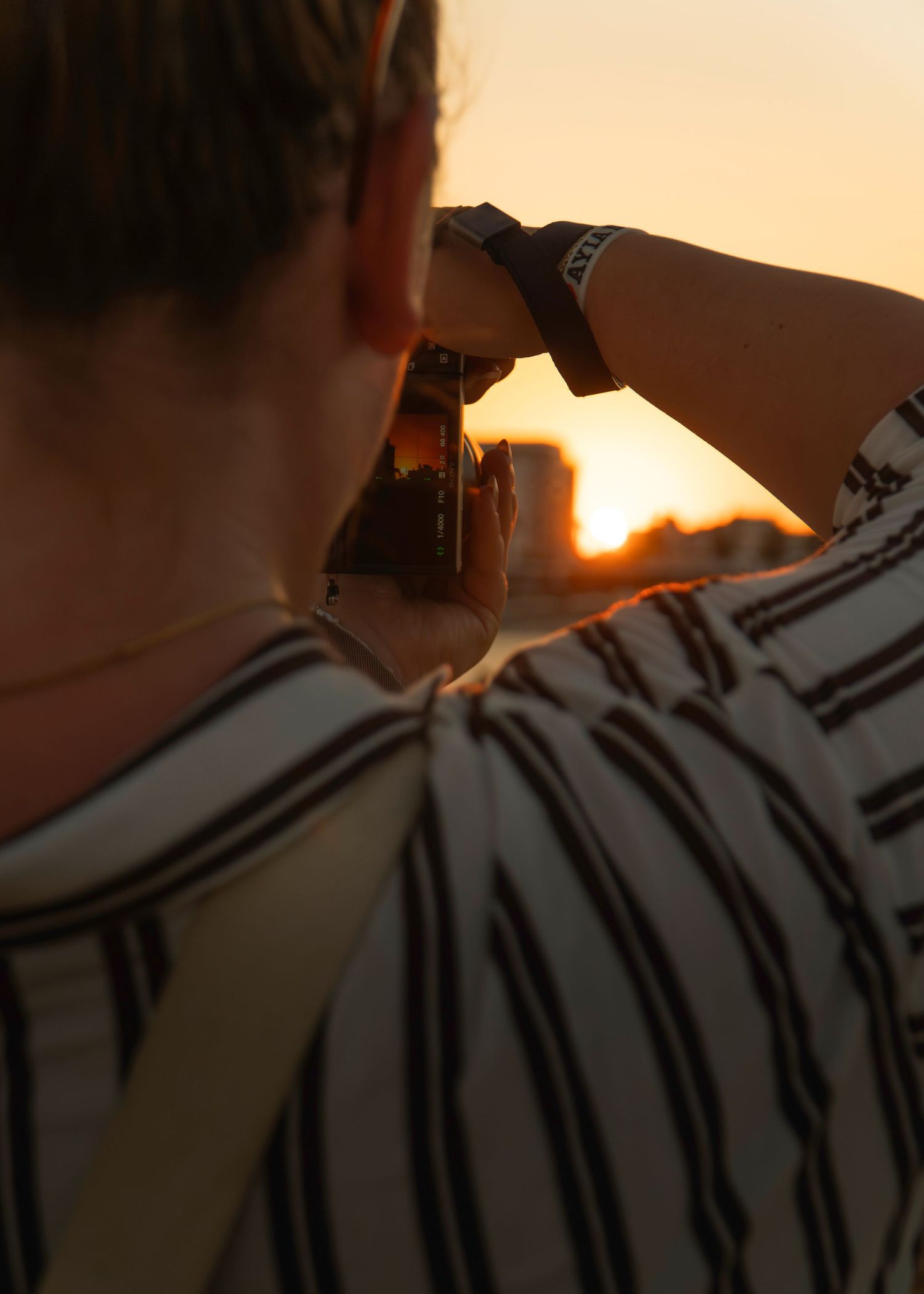Person photographing a golden sunset with a camera, wearing a striped shirt and black watch.