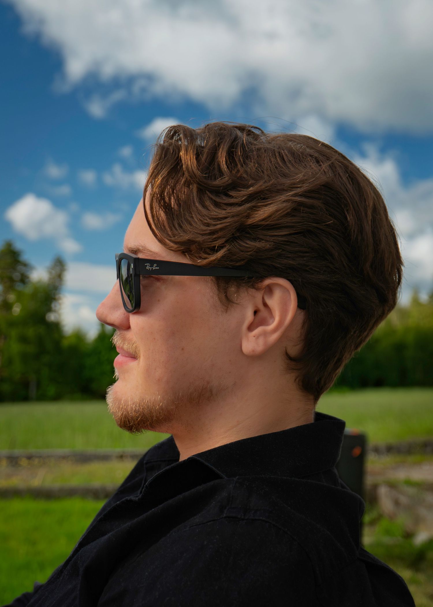 Young man in Ray-Ban sunglasses and black shirt photographed in profile against a blue sky backdrop.