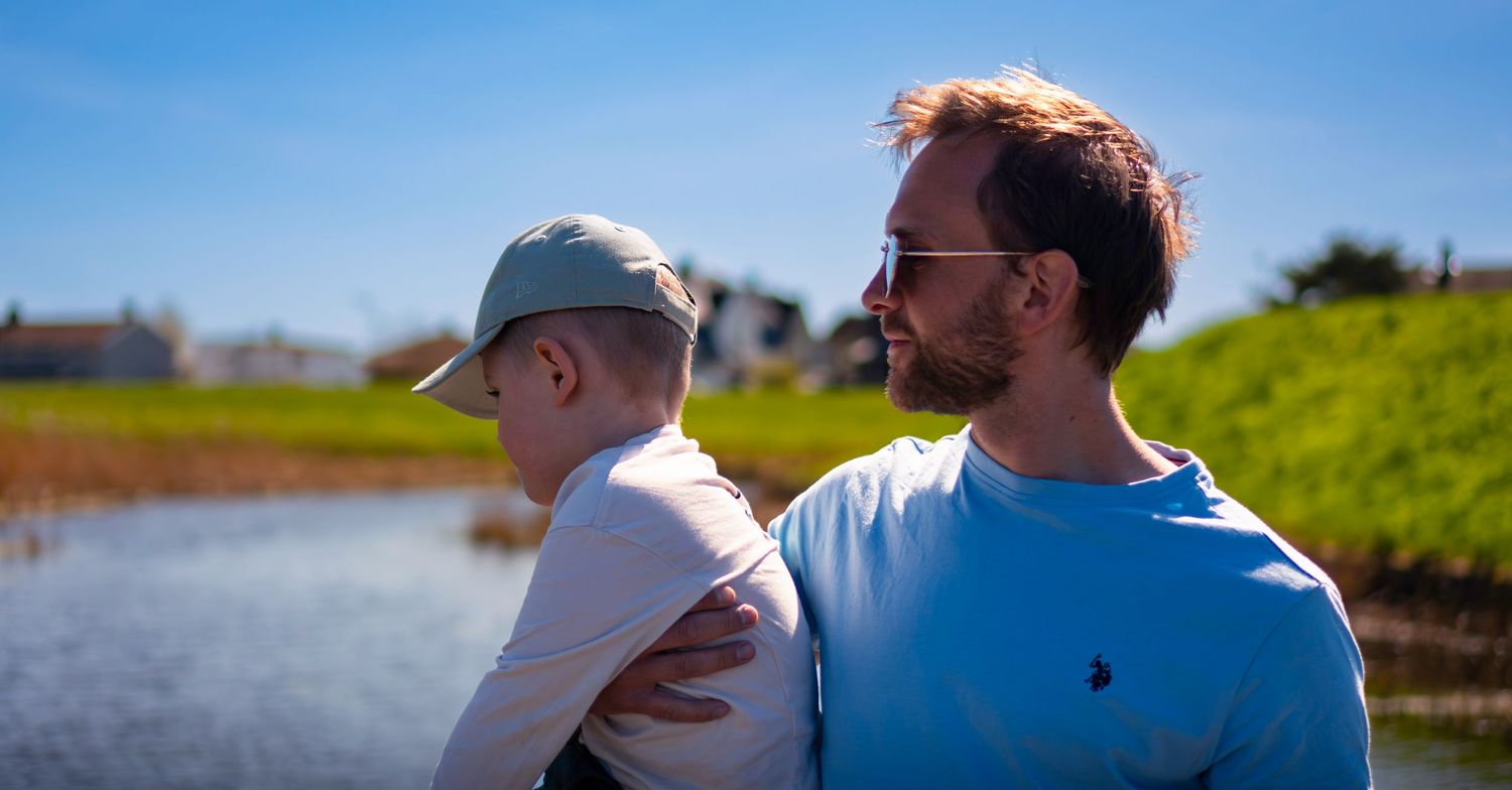 A man with sunglasses and beard stands with a child in a cap near a pond on a sunny day.