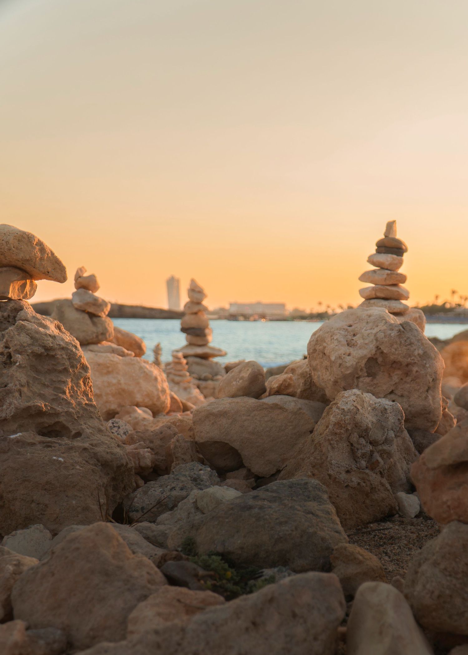 Stacked stone cairns on rocky beach at golden sunset with city skyline and calm water in background.