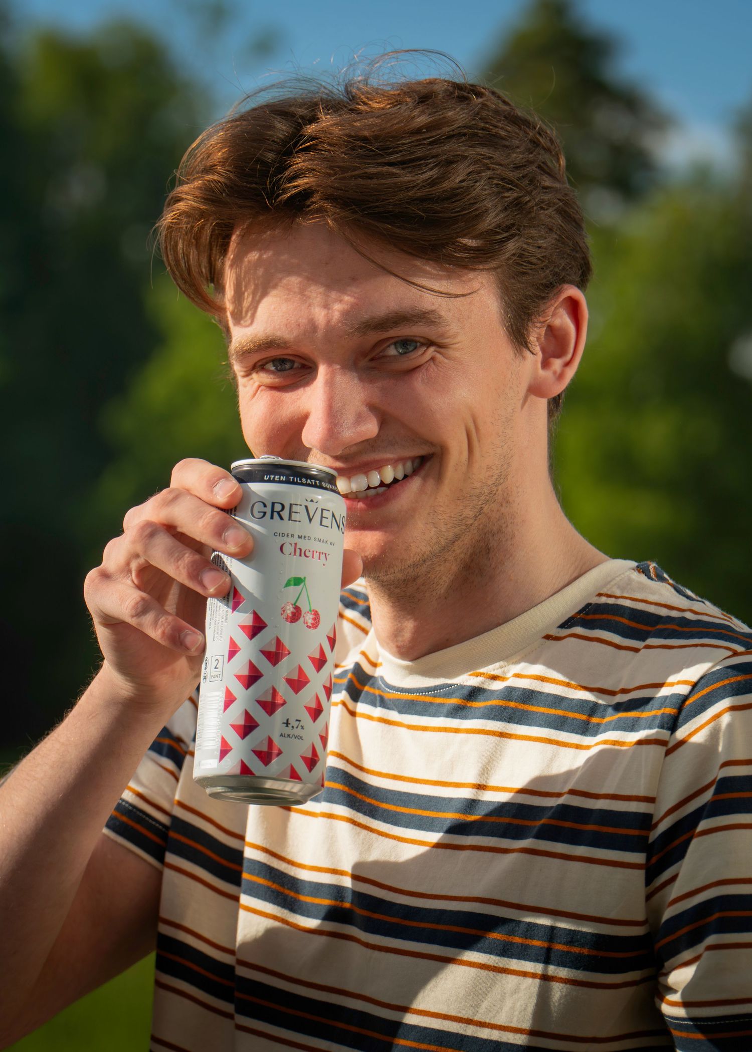 Smiling young man drinking a Greven Cherry sparkling water can outdoors in a striped shirt.