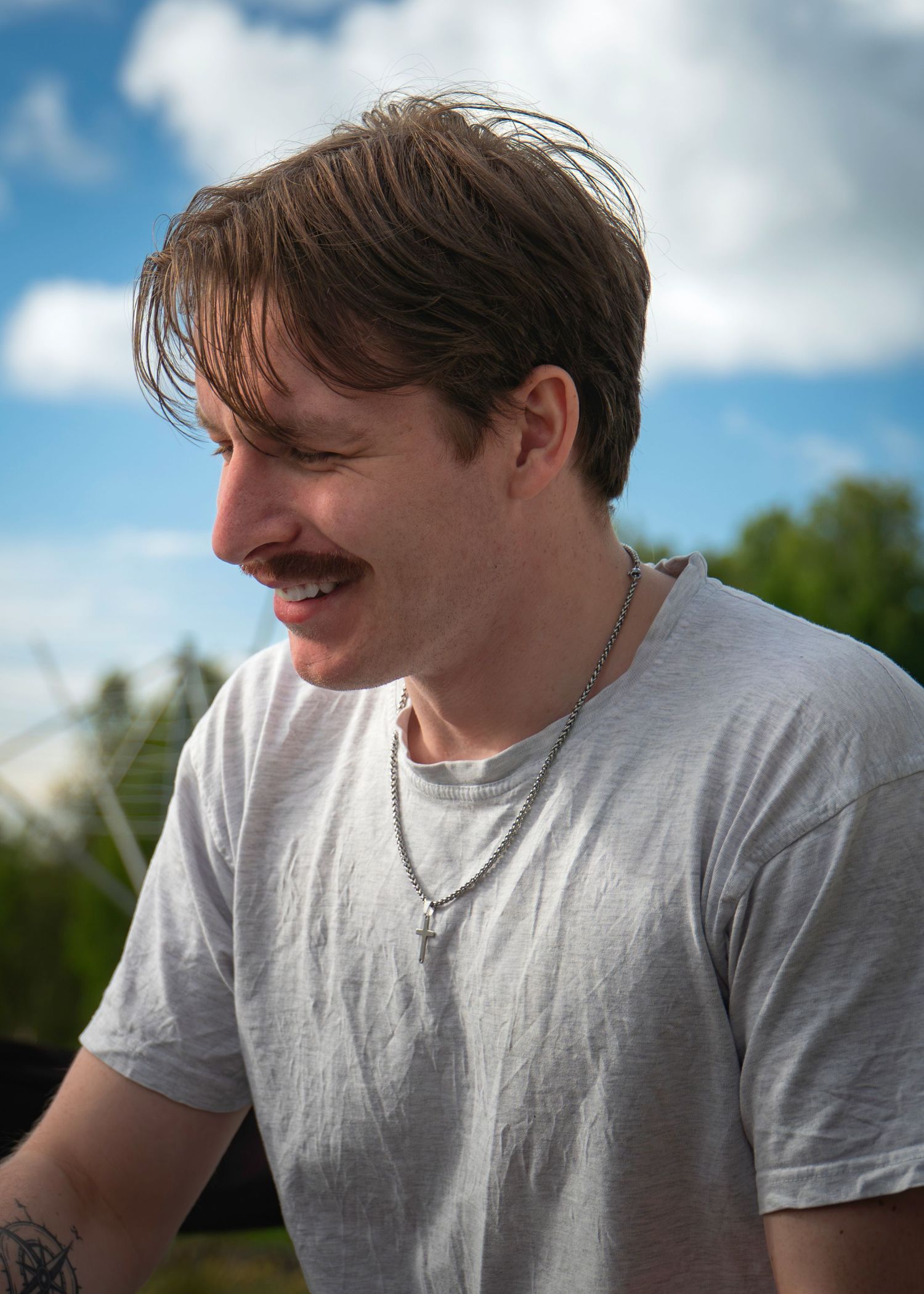 Young man with mustache smiling outdoors, wearing gray t-shirt and silver necklace against blue sky.