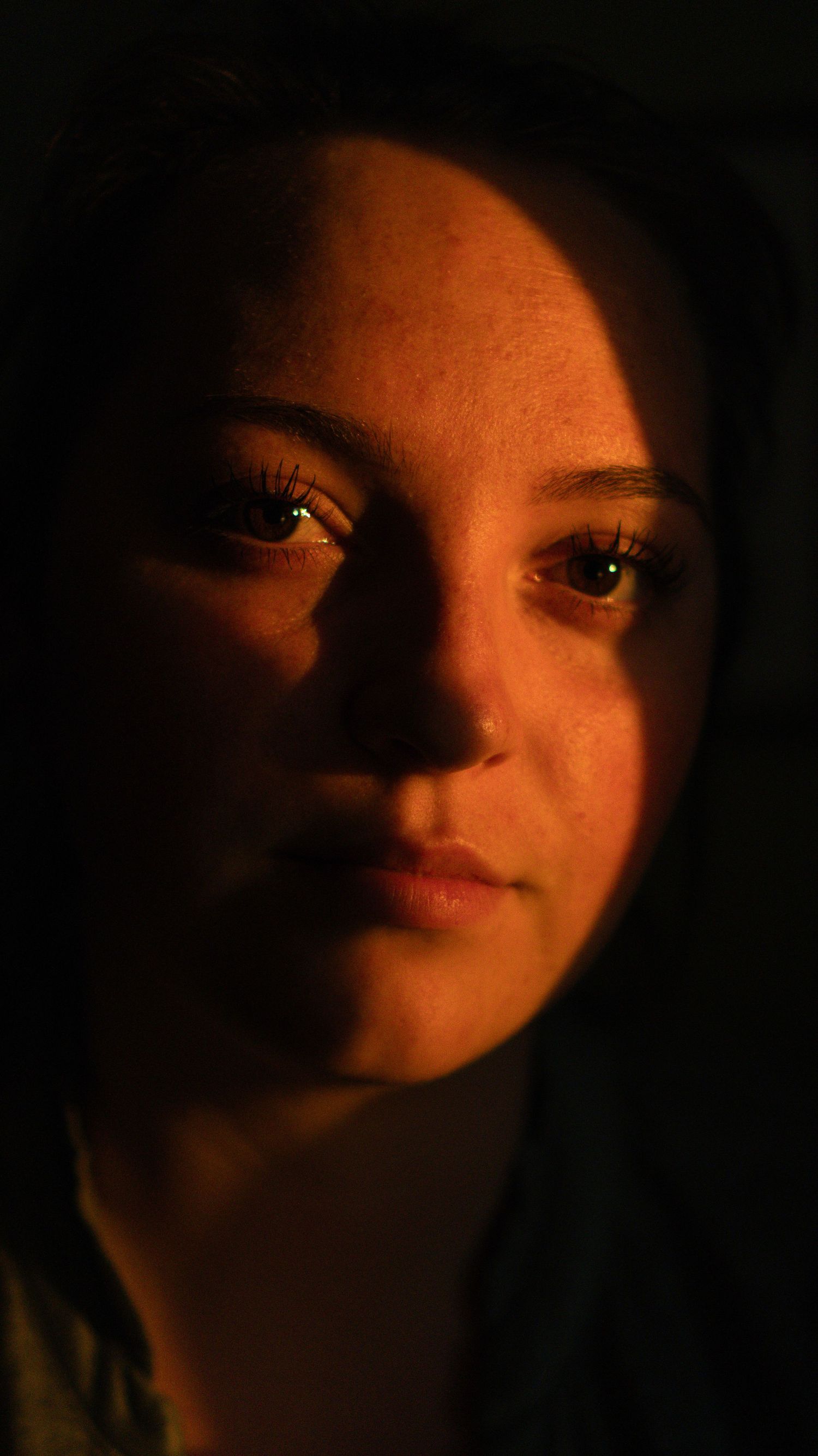 Close-up portrait of a young woman with hazel eyes illuminated by warm golden light against a dark background.