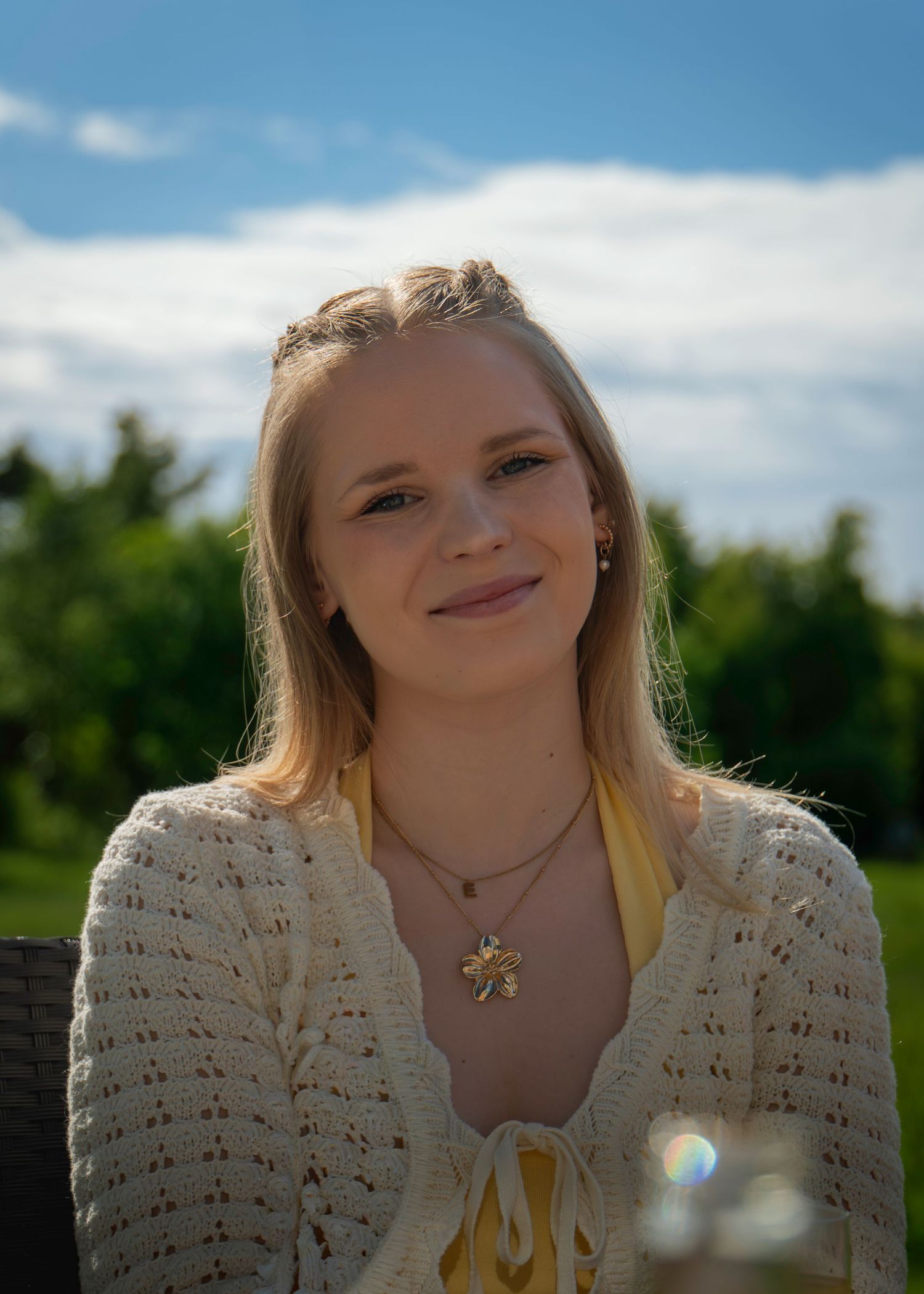 Young blonde woman smiling outdoors in cream knit cardigan and yellow top, with green trees and blue sky background.