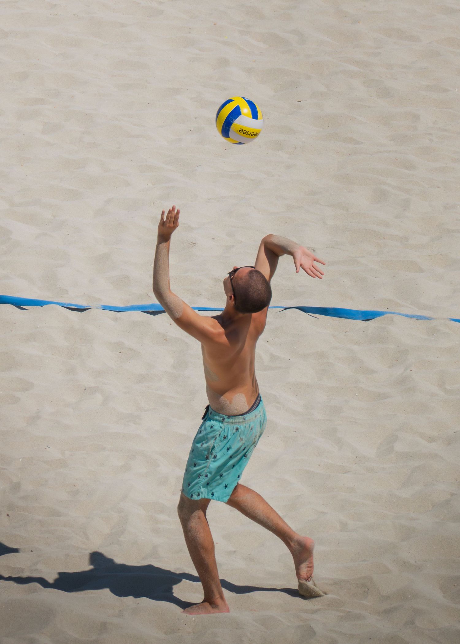 Shirtless man in teal shorts serving a volleyball on a sandy beach court with blue boundary line.