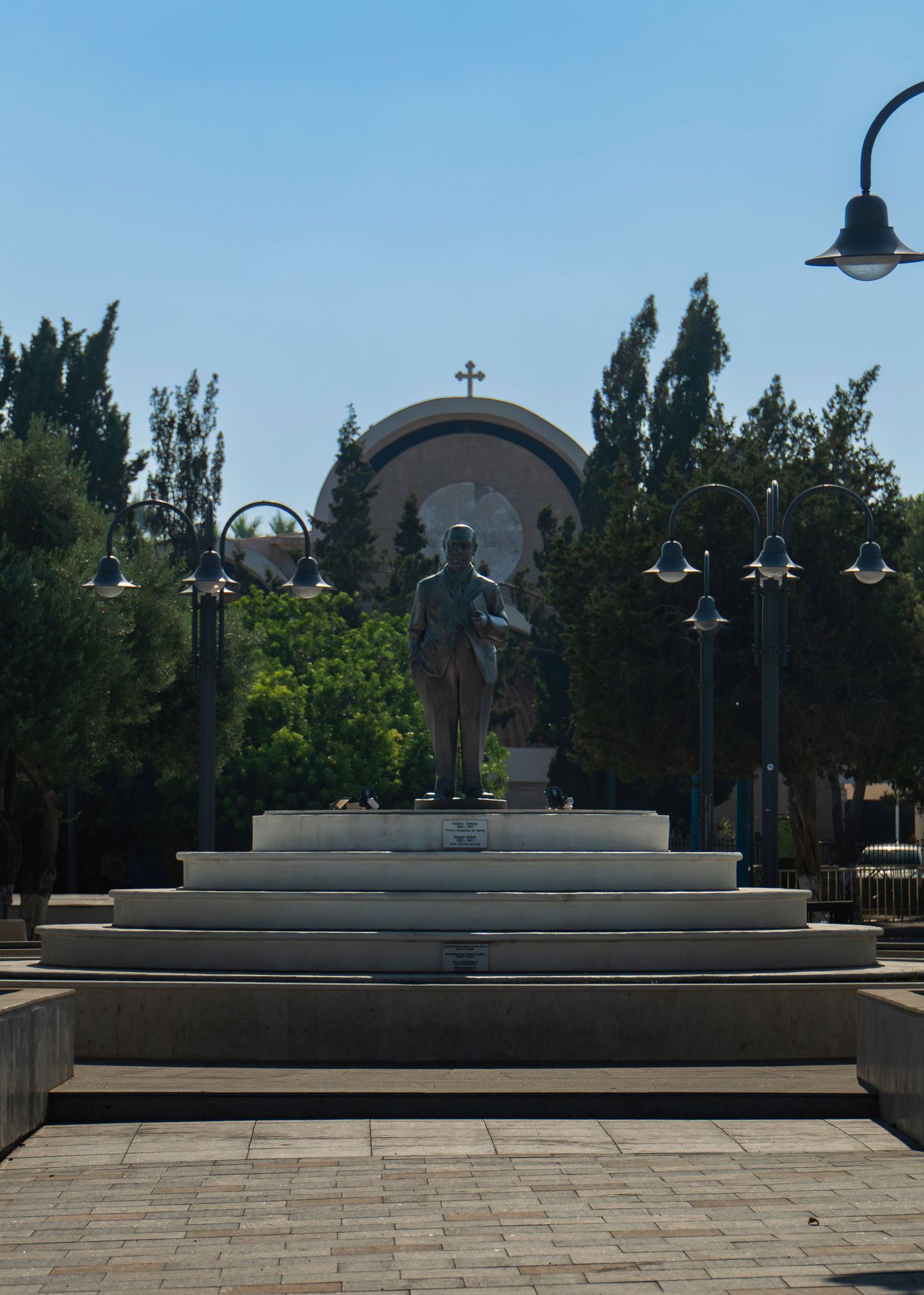 Bronze statue on tiered pedestal in a sunny plaza with street lamps, trees, and a domed church with cross in background.