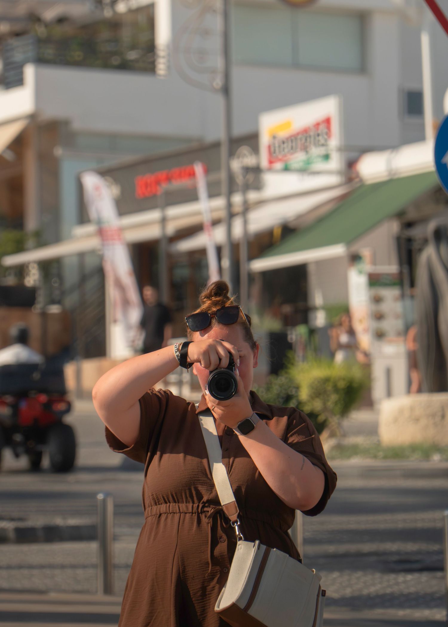 Woman in brown dress photographing with camera on busy street with shops in background on sunny day.