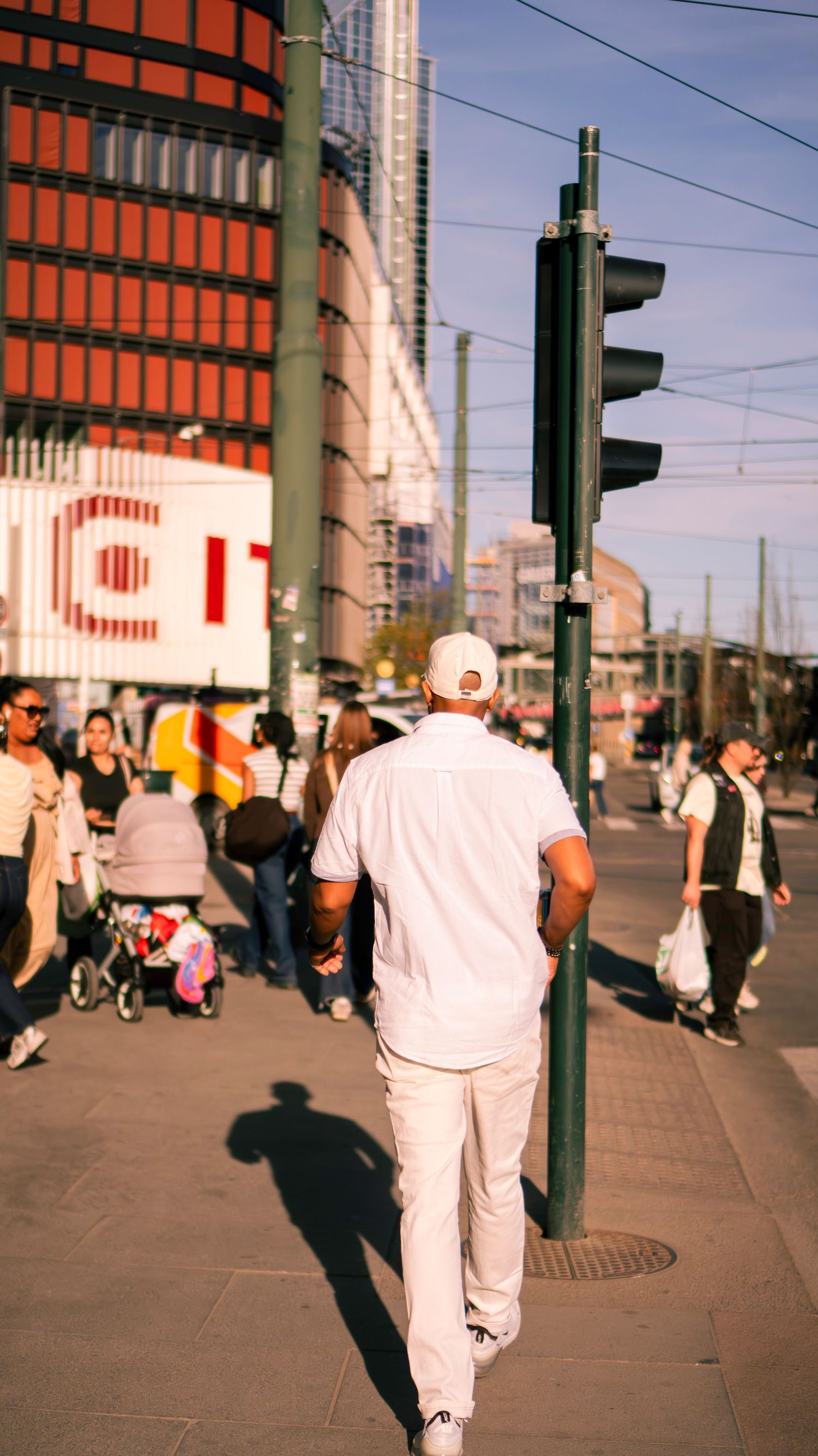 Man in white outfit walking on a busy city sidewalk near a traffic light, with pedestrians and urban buildings in the background.