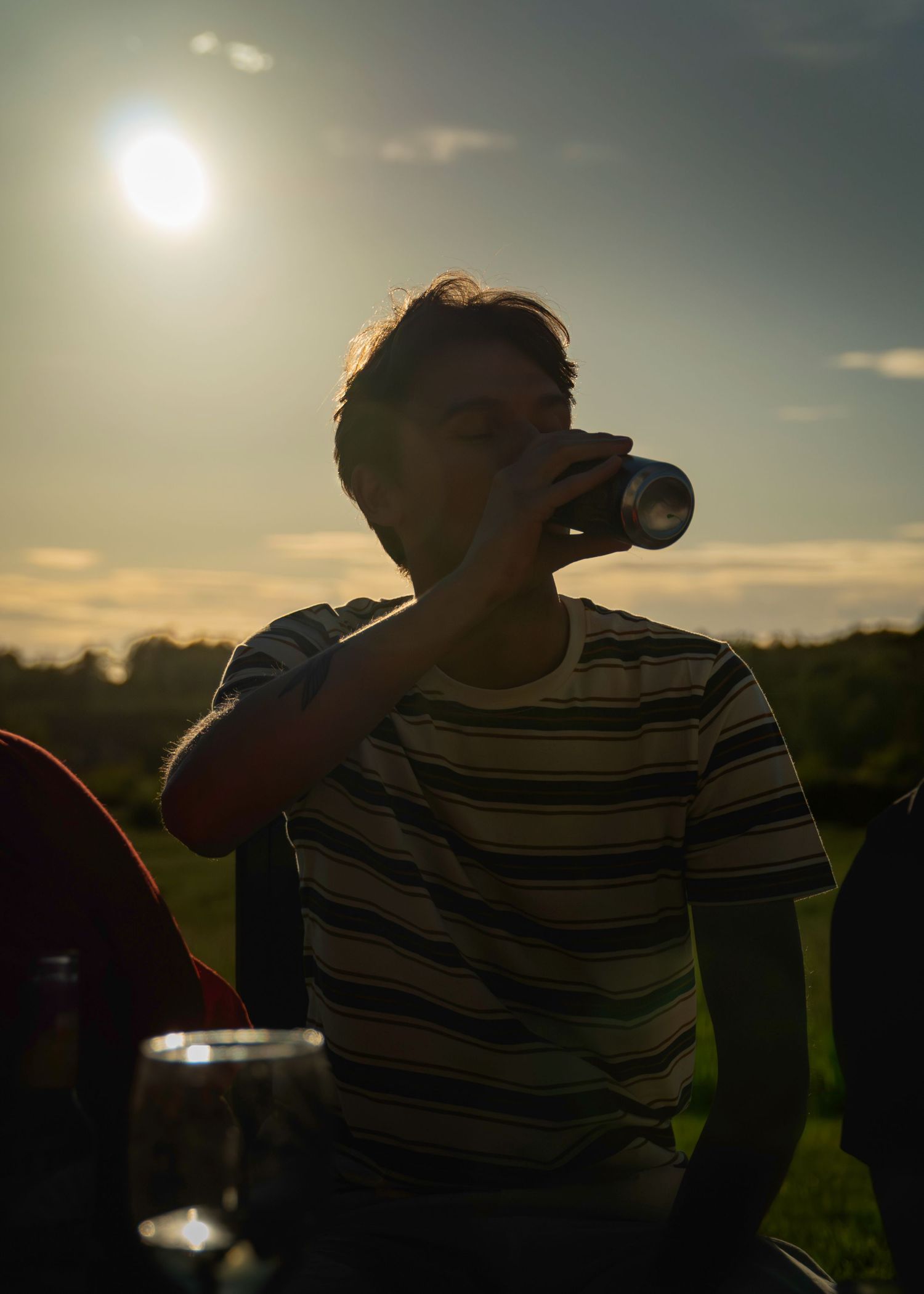 Young man in striped t-shirt drinking from a can outdoors at sunset with golden sunlight behind him.