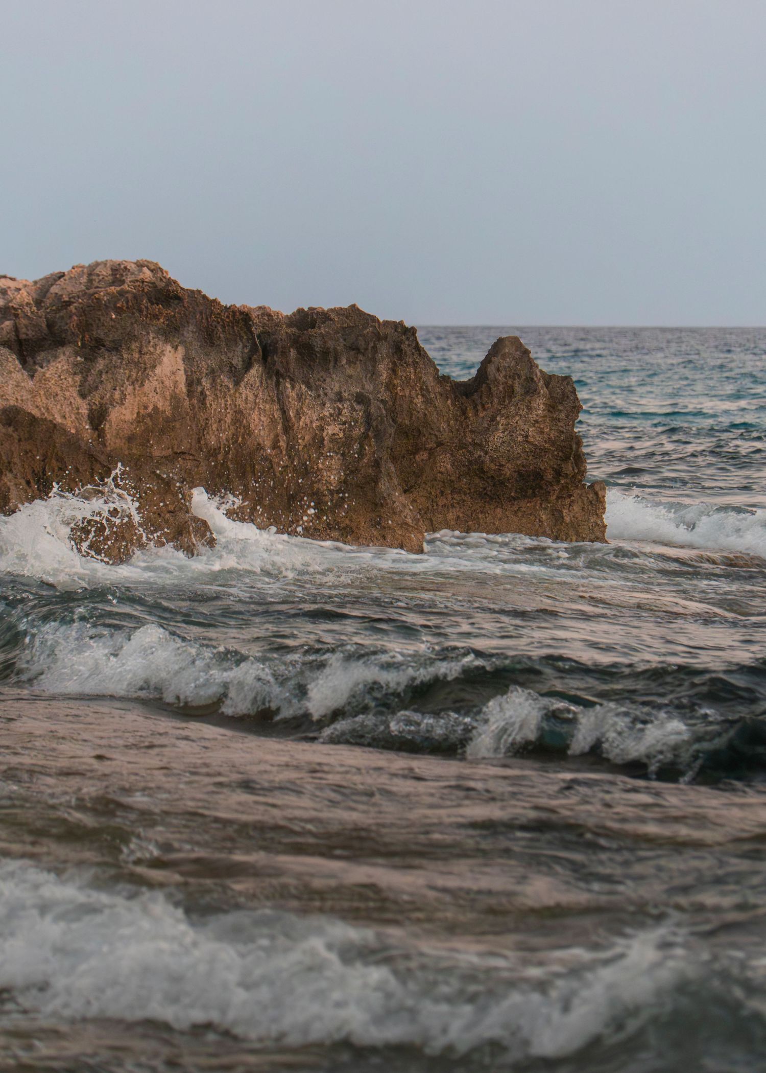 Waves crashing against rocky coastal cliffs with turquoise ocean water under an overcast sky.