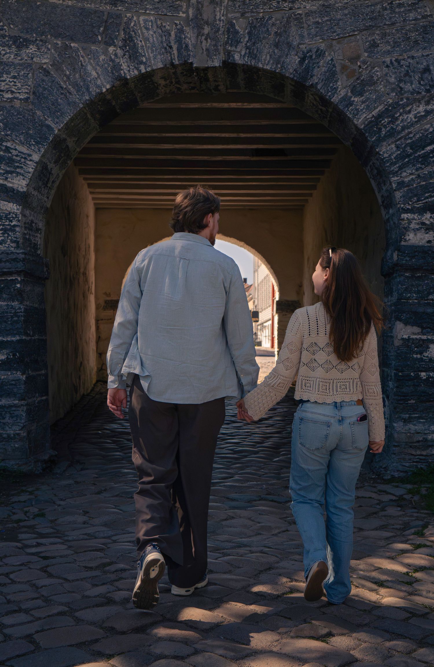 A couple holds hands walking through a stone archway tunnel on cobblestones in an old historic town.