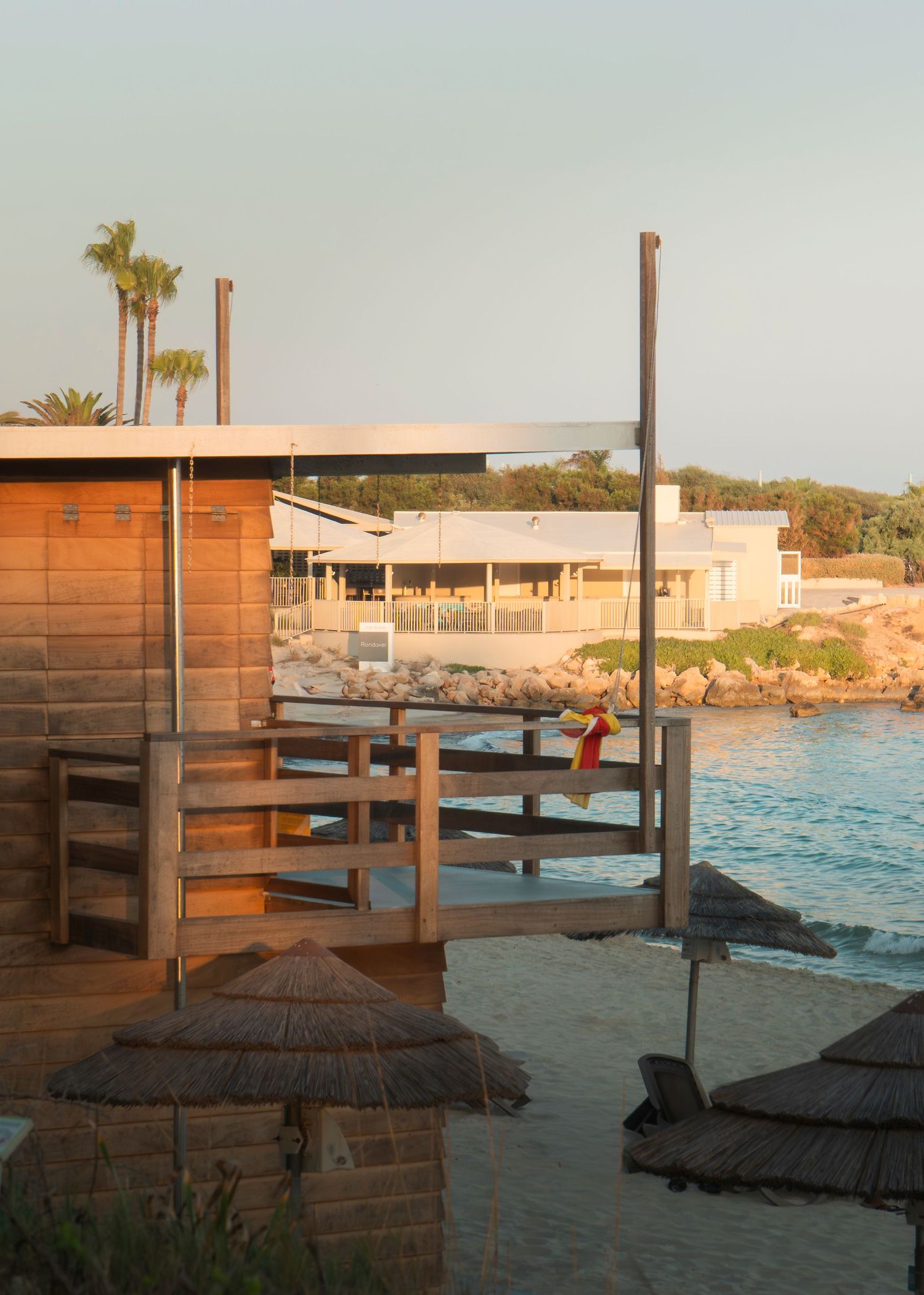 Beachside resort with wooden structures, thatched umbrellas, calm water, rocky shore, and palm trees at golden hour.