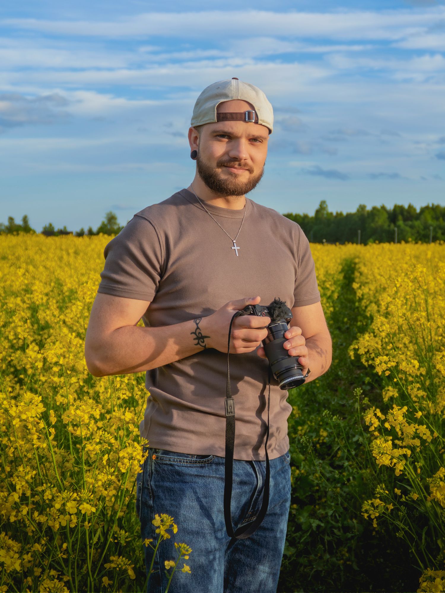 A muscular man in a gray t-shirt and cap holds a camera in a vibrant yellow rapeseed field under a blue sky.
