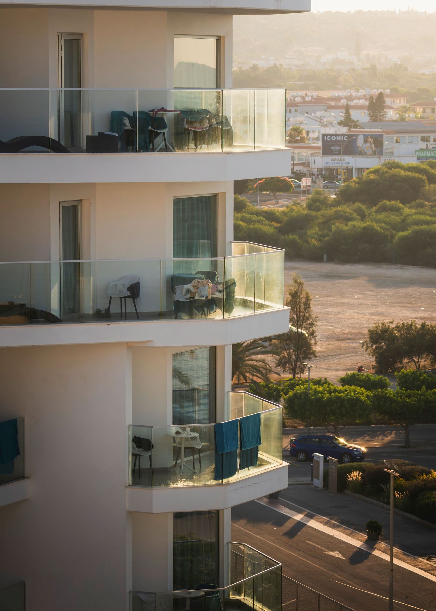 Modern white apartment building with glass balconies overlooking lush greenery and a coastal town at golden hour.