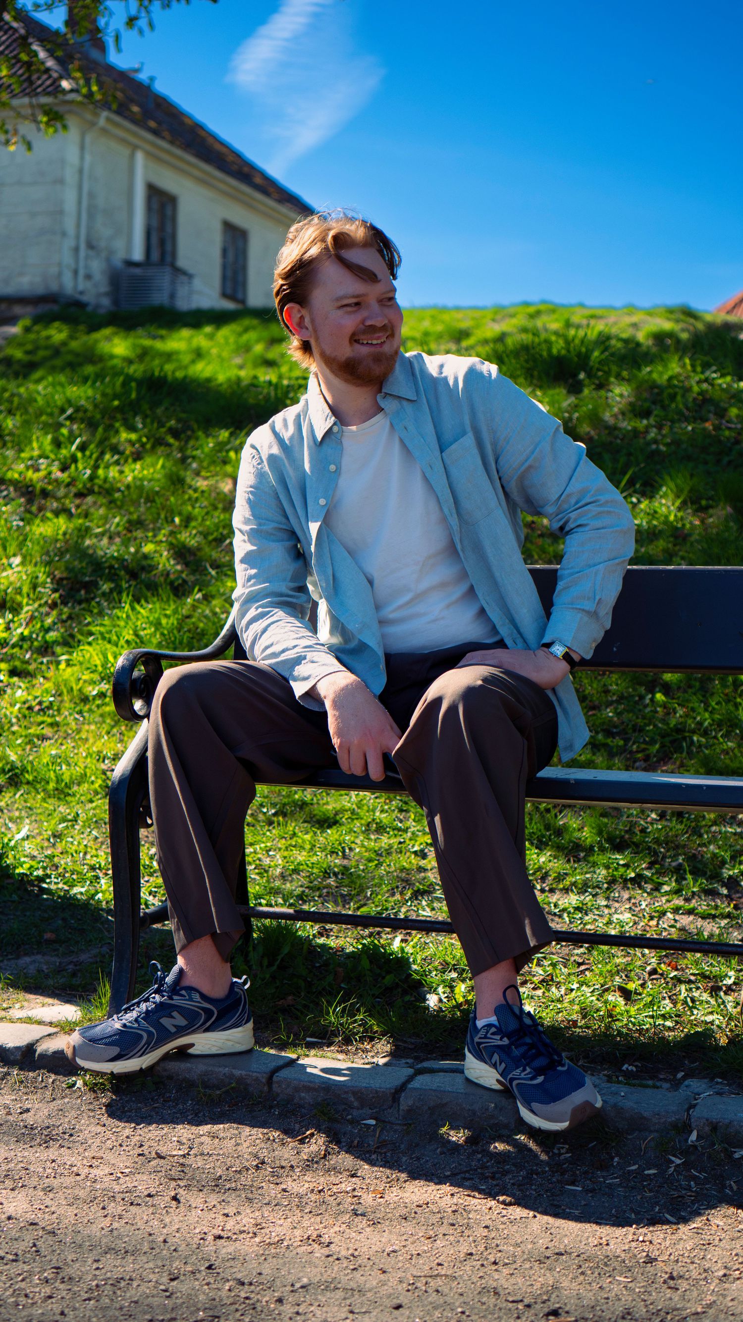 Young man with red hair sitting on a park bench outdoors, wearing a light blue shirt and brown trousers on a sunny day.
