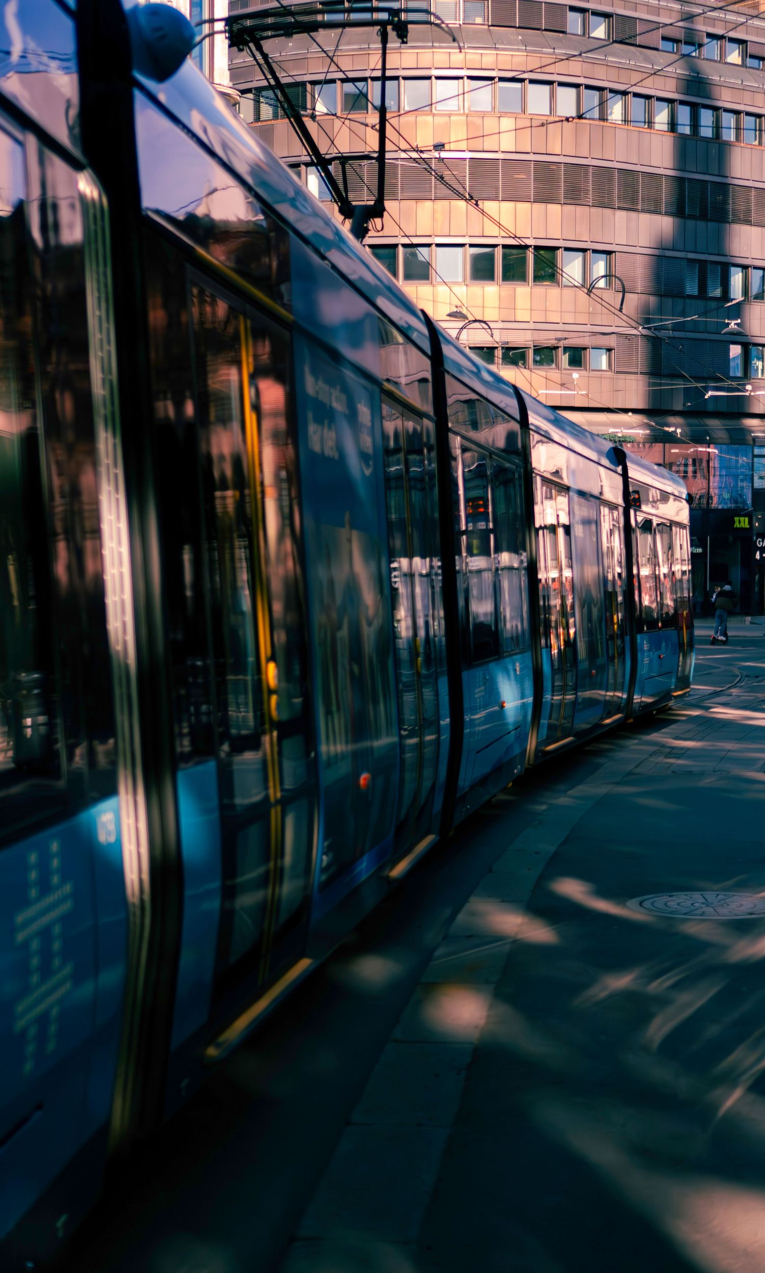 A modern blue tram glides through an urban street, reflecting city buildings in its windows on a sunny day.