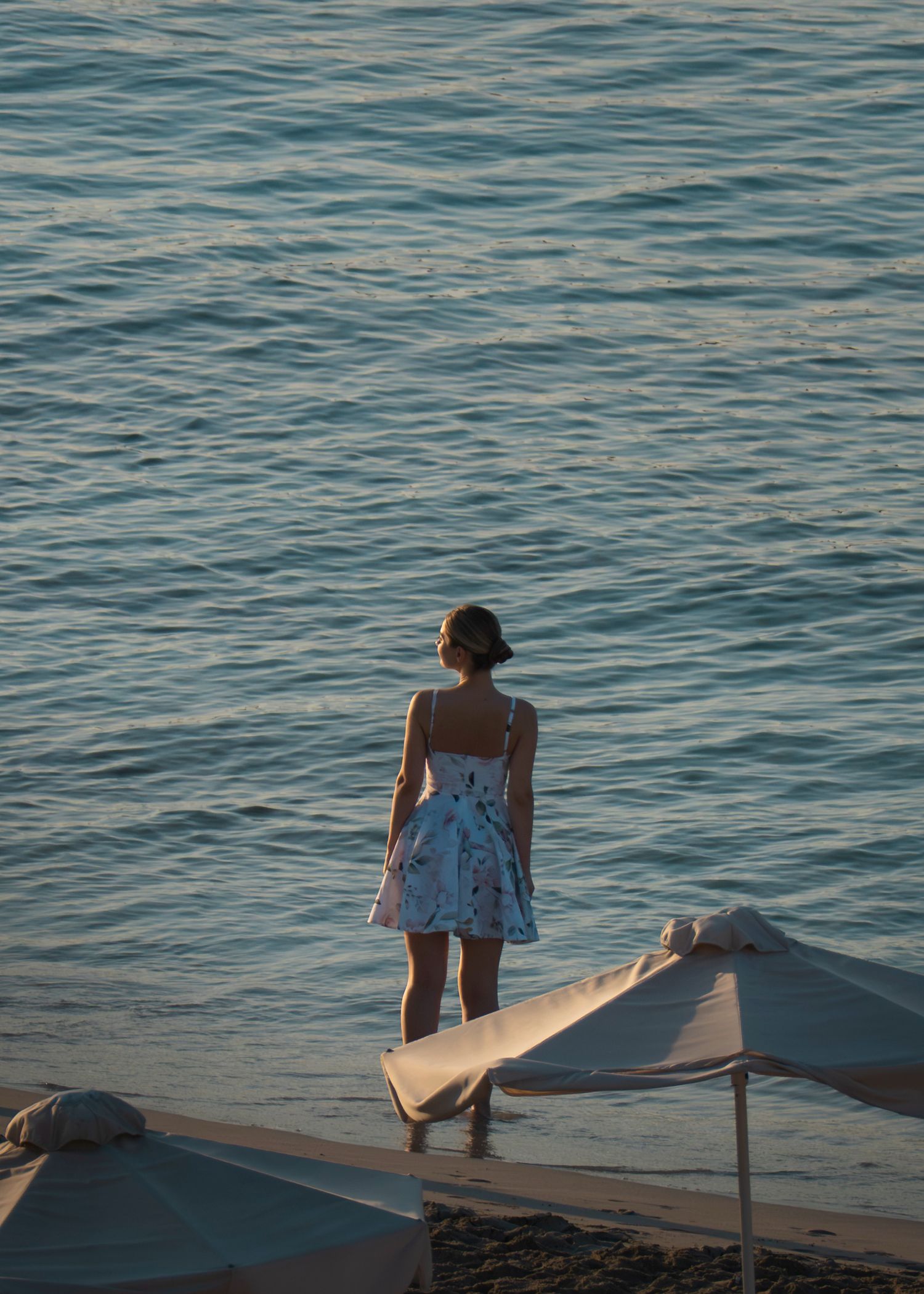 Woman in white floral dress standing on beach shore gazing at calm blue ocean waters at dusk.