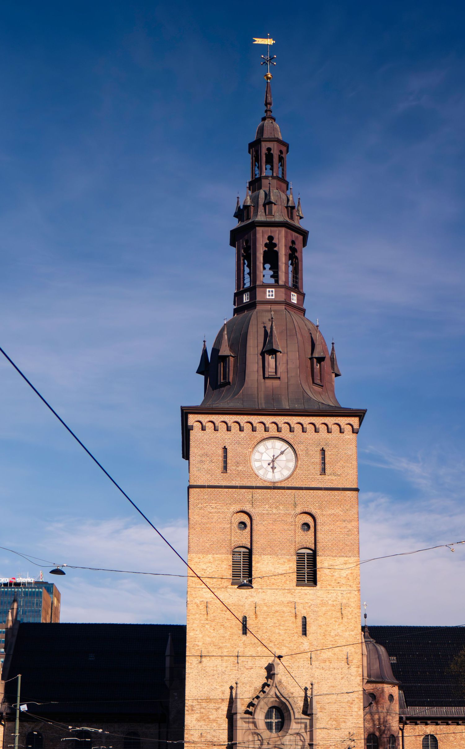 Historic brick church tower with clock face and ornate copper spire against blue sky in Oslo, Norway.