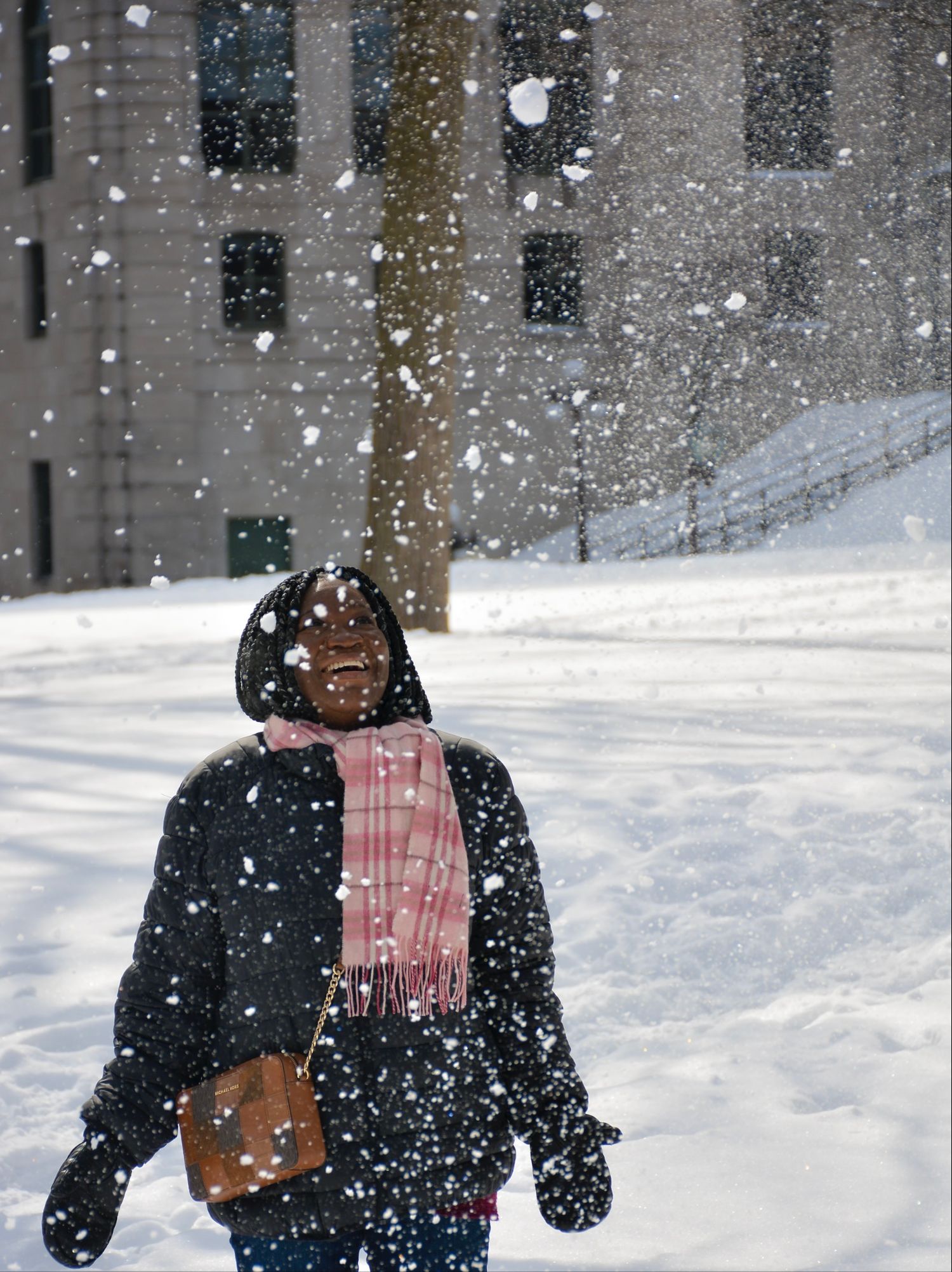 Woman standing outdoors in the snow, smiling with arms outstretched, wearing a pink scarf and winter coat