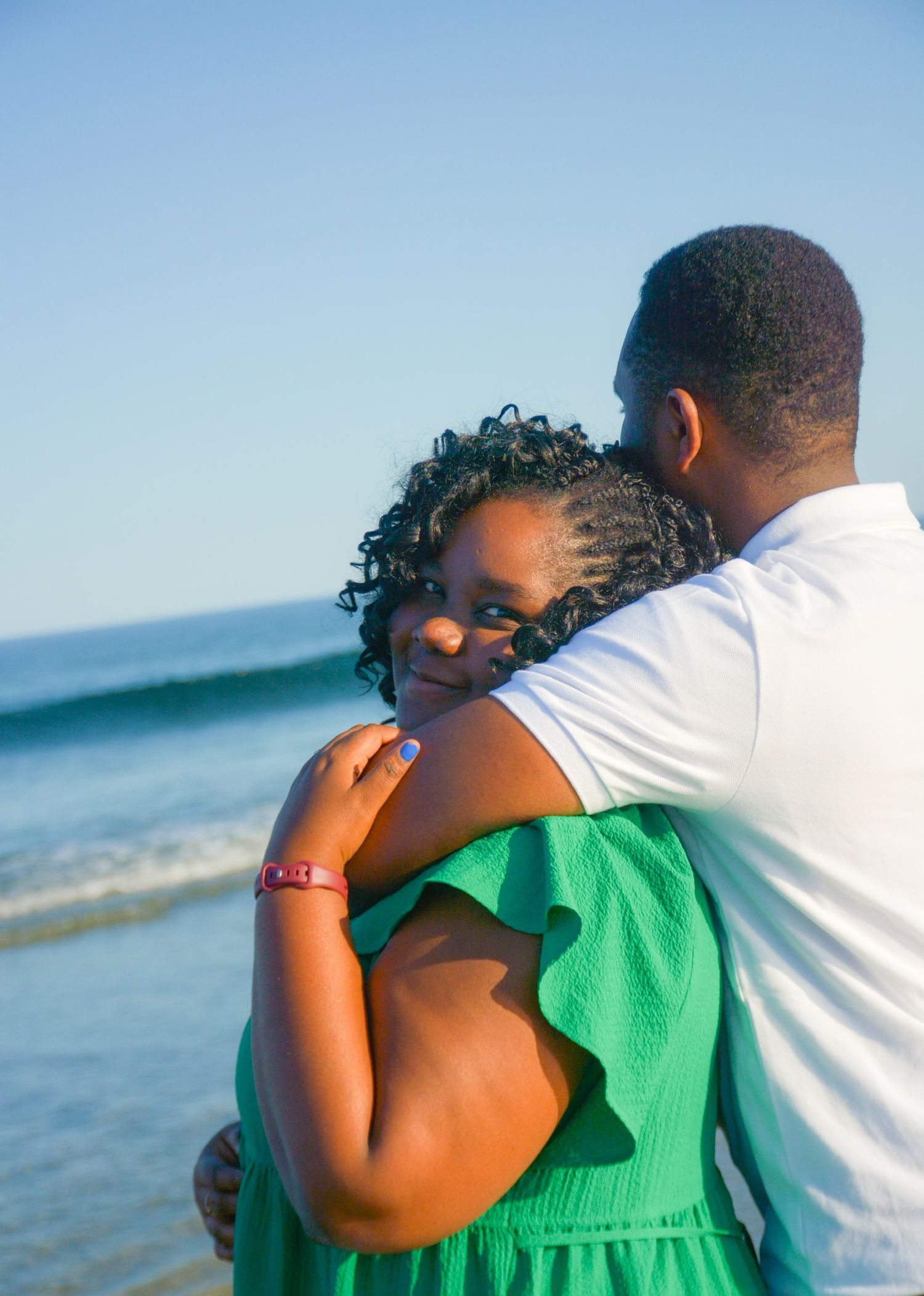 Couple embracing on the beach, woman in a green dress resting her head on her partner's shoulder with the ocean behind them
