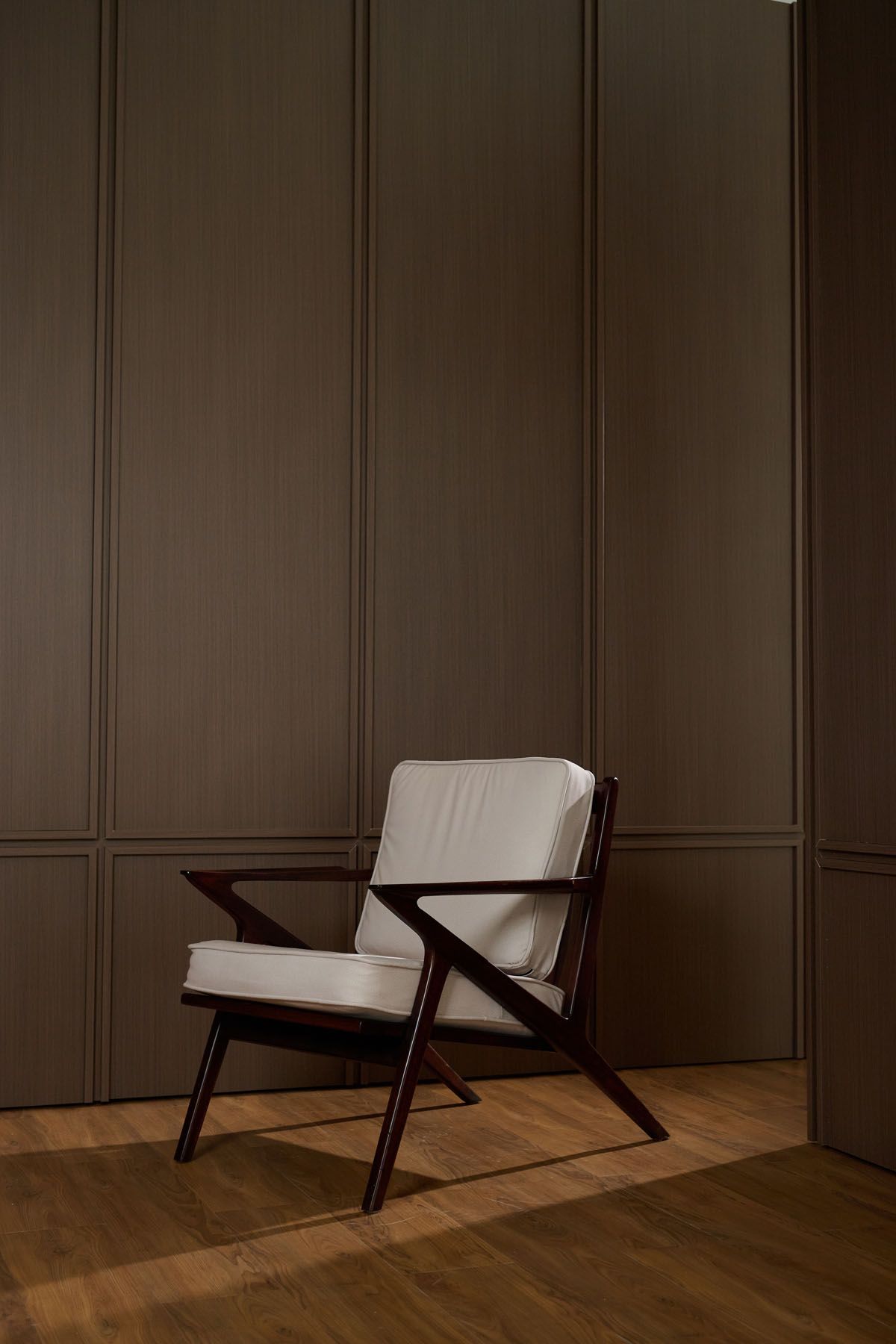 Mid-century modern armchair with white cushions and dark wood frame against brown paneled wall on hardwood floor.