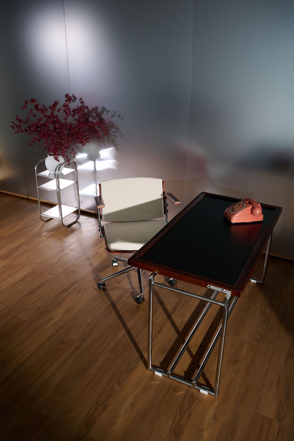 Modern office with chrome desk, pink rotary phone, white chair on wheels, and red branch decor on hardwood floor.