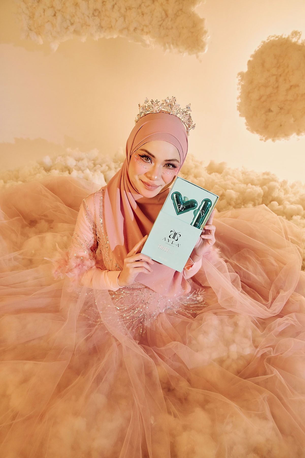 Muslim woman in pink hijab and tiara crown holds Ayla Cosmetics Twist lip gloss gift set.