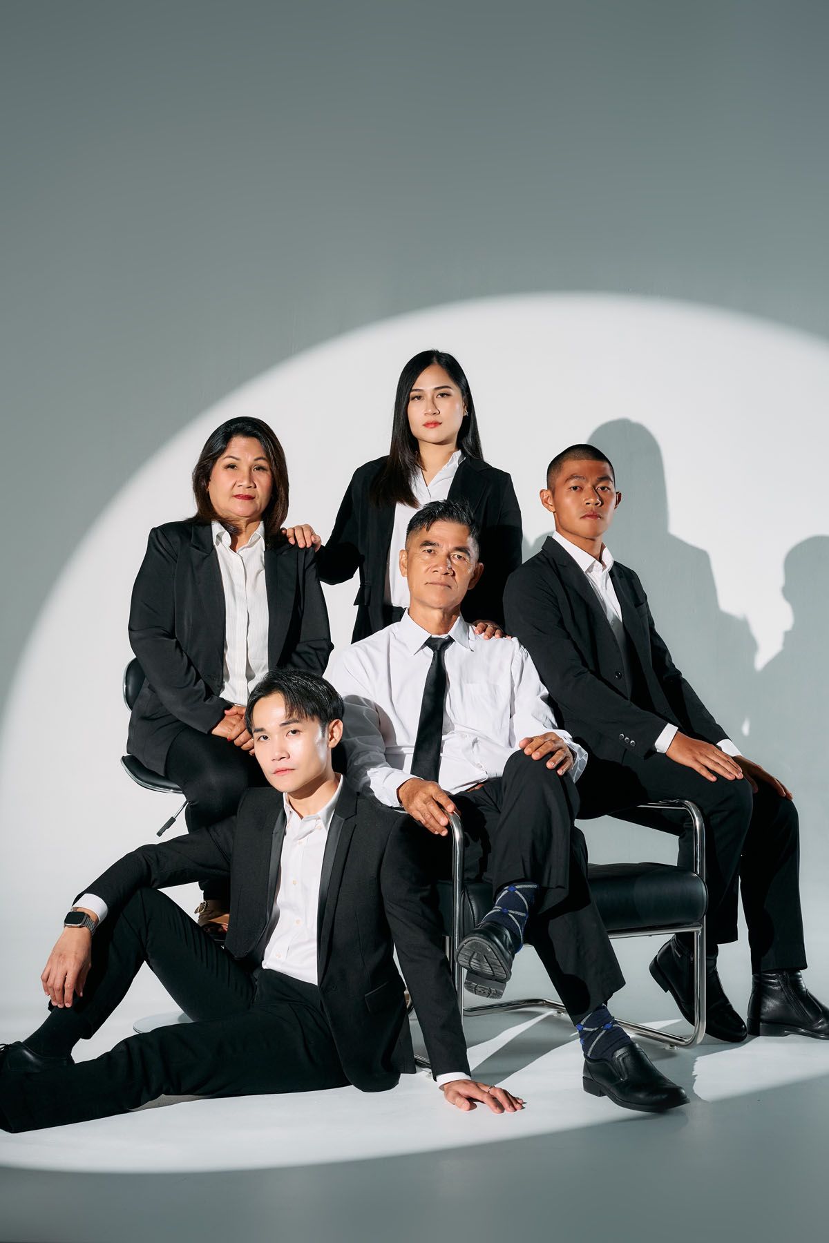 Five Asian professionals in black suits pose confidently under a spotlight against a gray background.